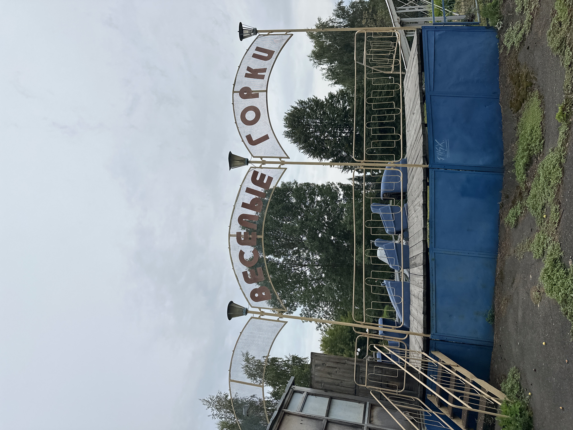The image shows a dilapidated amusement park ride in Biysk, Russia. The ride is a roller coaster with blue seats on a wooden platform. A sign above the ride reads "Веселые Горки," which translates to "Merry Hills." The surrounding area is overgrown with weeds, and the structures appear to be in disrepair. The sky is overcast.