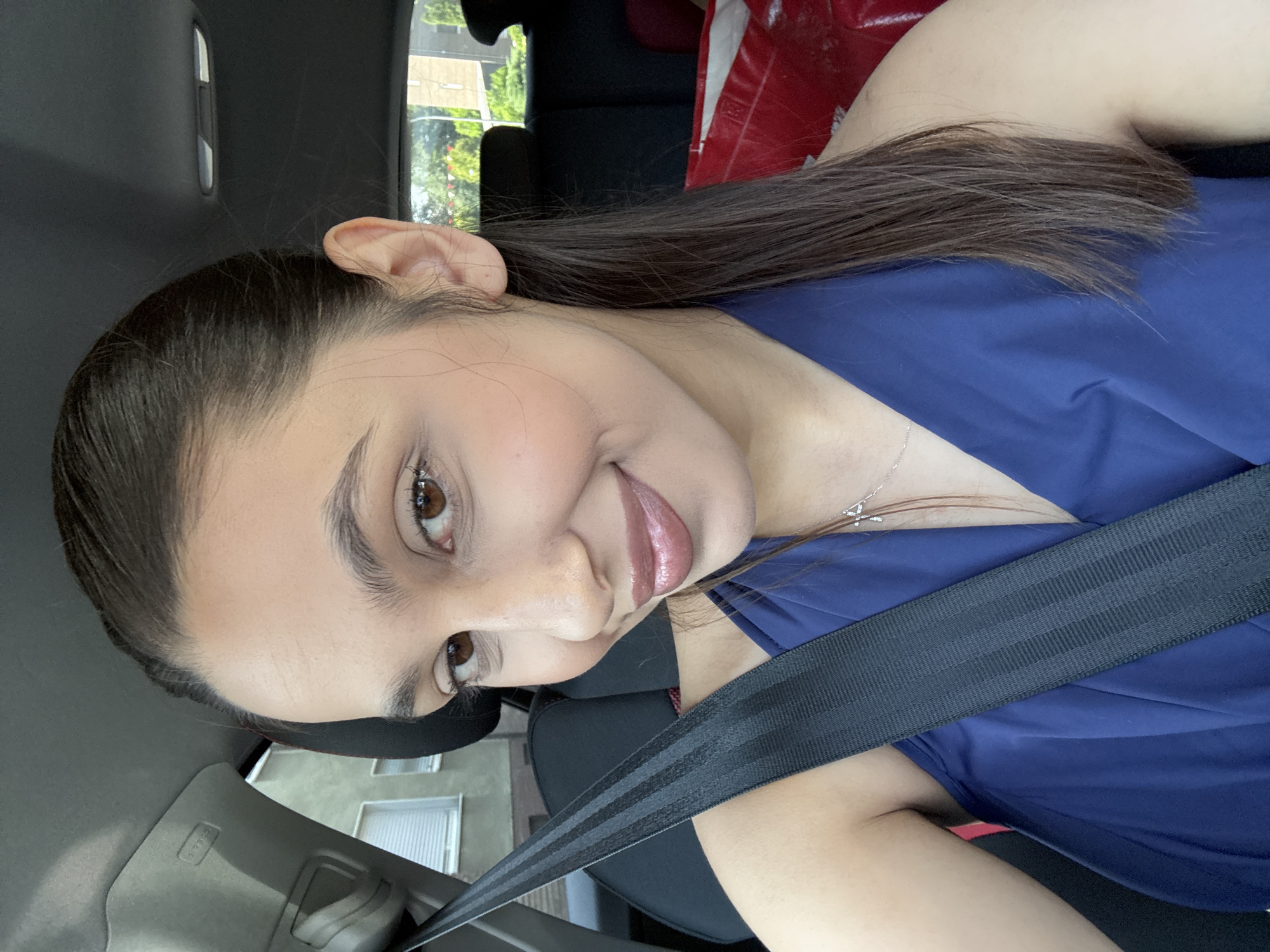 A young woman is seated in the driver's seat of a car. She is wearing a blue top and a seatbelt. She has long, dark hair pulled back in a ponytail. Her facial features are visible. The background shows a glimpse of a street with buildings. The image may have been taken in Mönchengladbach, Germany.