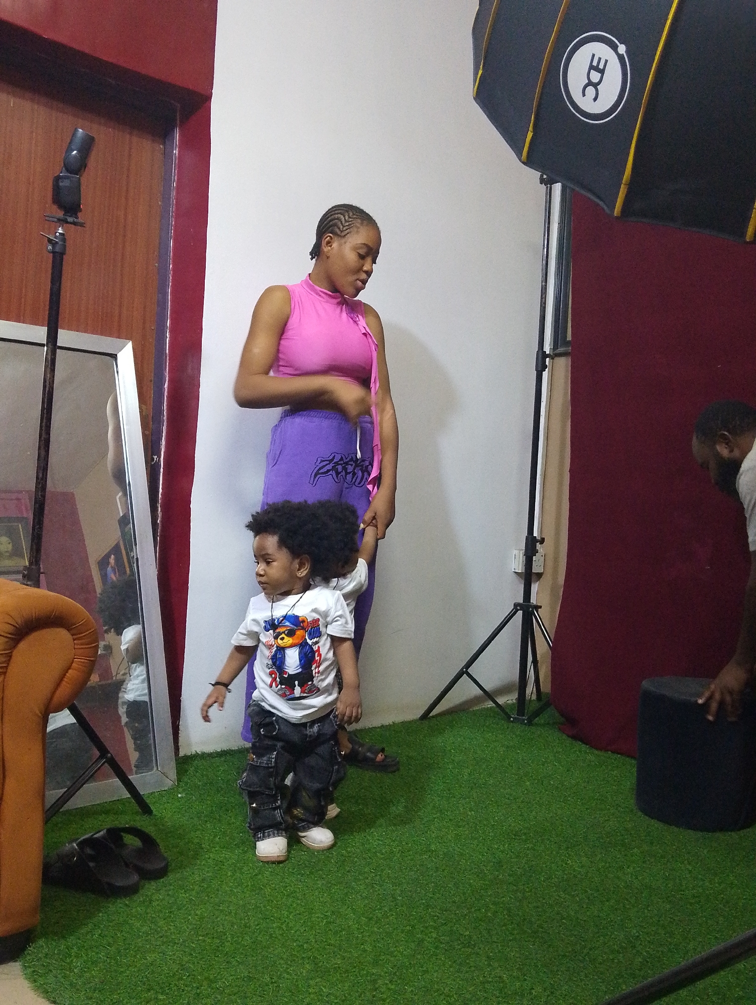 This indoor full-length shot captures a scene within what appears to be a photography studio in Babban Giji, Nigeria. The floor is covered with artificial green turf, while the background consists of a plain white wall on the left and a dark red wall or backdrop on the right.
In the center-left, a young woman with braided hair stands, wearing a sleeveless pink top and purple pants. Her gaze is directed downwards, and she appears to be holding the hand of a young child beside her. The child, likely a boy, has dark curly hair and is dressed in a white t-shirt featuring a colorful cartoon bear graphic, dark cargo pants, and white shoes. He looks towards the left with a curious or focused expression.
Photography equipment is prominent, indicating a professional setup. A speedlight flash on a stand is positioned on the far left. To the right, a large black softbox with a white "EDC" logo (encircled) is mounted on another stand. Further right, a partial view of a man with a beard, wearing a light-colored t-shirt, is seen bent over a black cylindrical stool or ottoman, seemingly preparing or adjusting equipment.
Additional objects include a large, silver-framed full-length mirror on the left, reflecting portions of the room and the child. An orange upholstered armchair is partially visible on the far left. A pair of black slide sandals rests on the green turf near the mirror. A power outlet is visible on the white wall near one of the light stands. The scene suggests a photoshoot is either underway or being set up.
