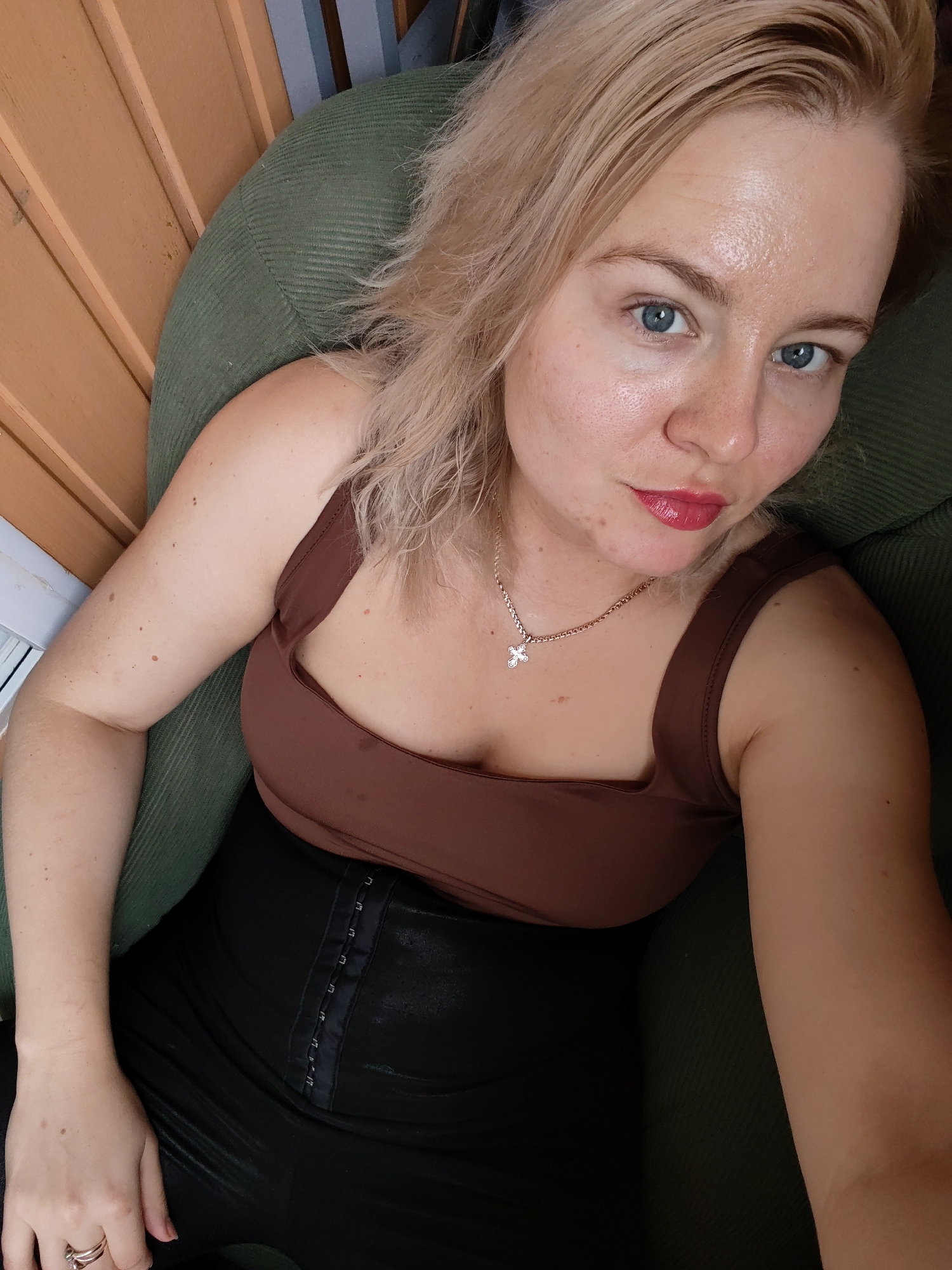 A woman with blonde hair and blue eyes sits in a green chair. She is wearing a brown tank top and black leggings. She has a cross necklace around her neck and a ring on her left hand. The background appears to be indoors, with wooden paneling visible. There is no specific information available to determine if this image was taken in Izhevsk, Russia.
