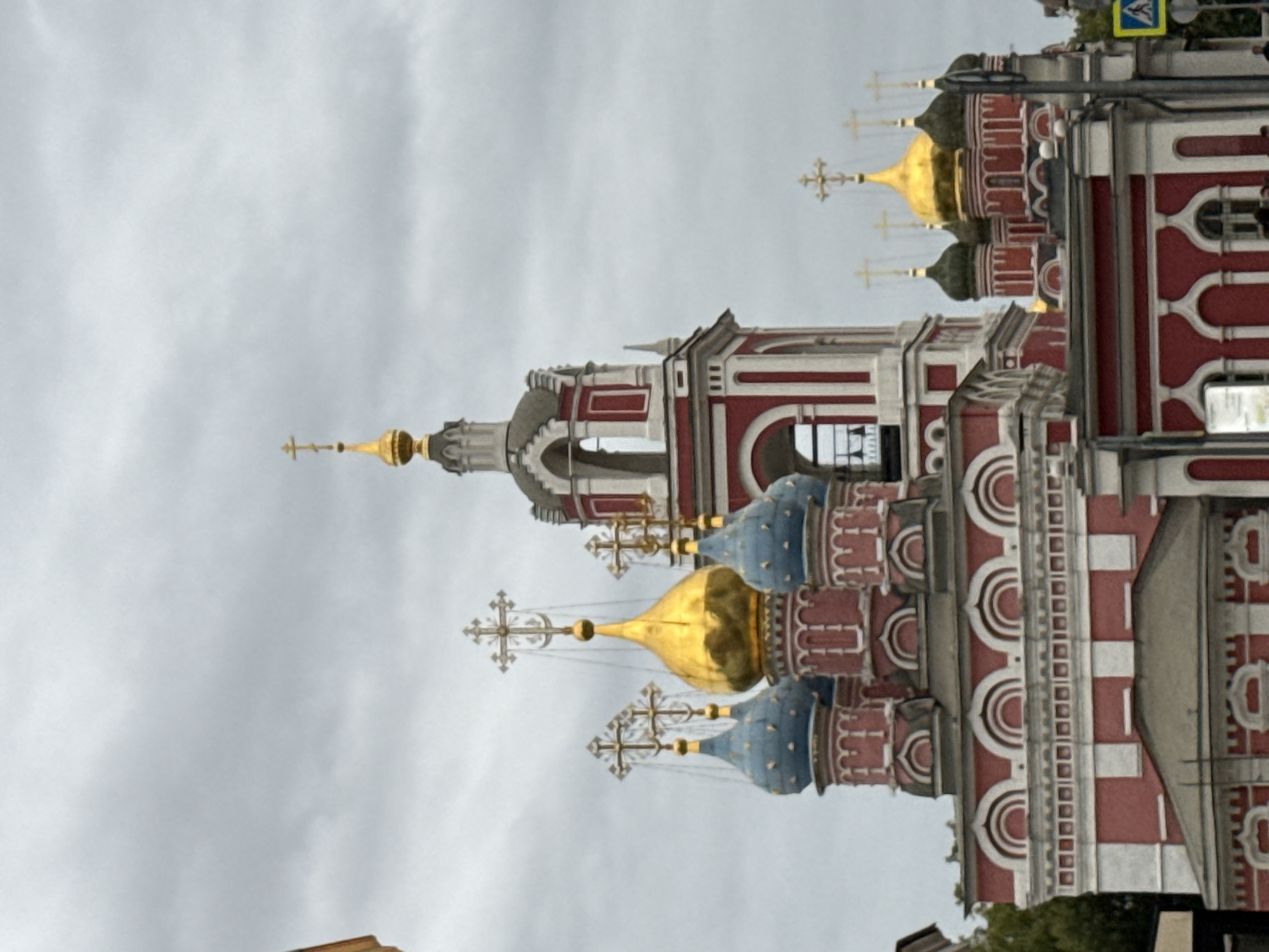 The image captures the exterior of the Saint Basil's Cathedral, a prominent landmark located in Moscow, Russia. The cathedral is characterized by its vibrant, onion-shaped domes, some of which are gilded and others painted in blue and red. The architecture features intricate details with red and white striped patterns. The sky above is overcast with gray clouds, suggesting a cloudy day. The foreground shows a portion of the cathedral's facade, with its distinct red and white coloring and arched windows. In the lower right corner, a yellow traffic sign with a blue pedestrian symbol is visible. The image appears to be taken from a street level perspective, with a slightly upward angle.