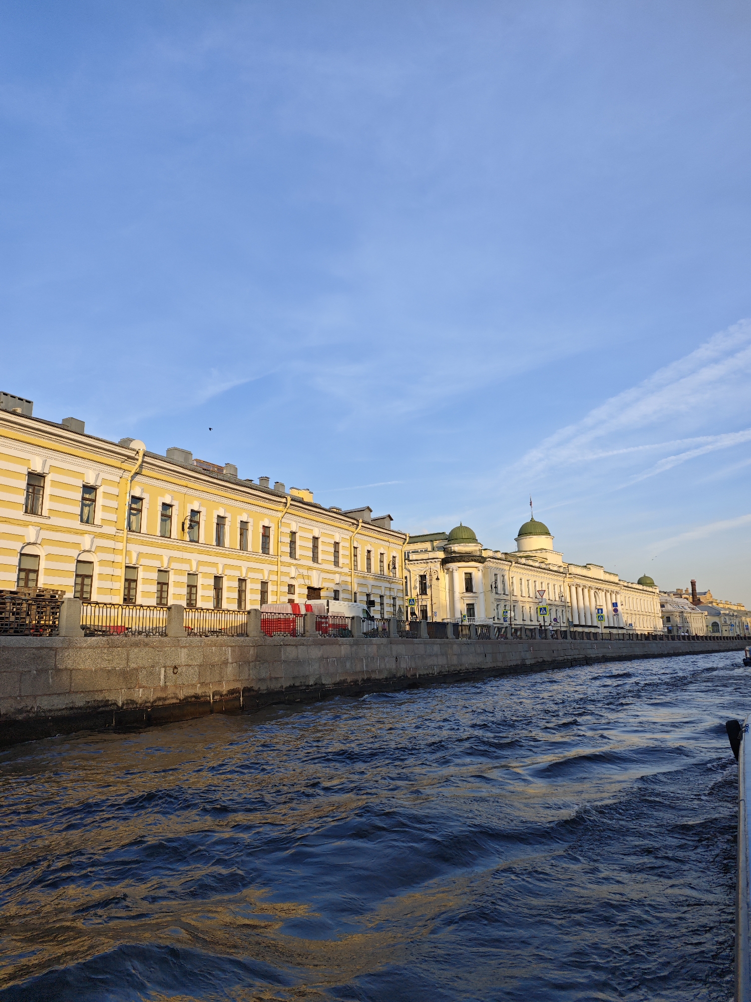 The image shows a wide, deep canal with dark blue, rippling water reflecting golden sunlight and the blue sky. Along the canal's edge, a stone embankment supports a row of buildings. The buildings are predominantly white with yellow accents, featuring classical architectural elements like columns and green domes on some structures. The first building on the left is yellow with white horizontal stripes and numerous windows. People are visible walking along the embankment and crossing streets in front of the buildings. Traffic signs and streetlights are also present. The sky is a clear, light blue with sparse, wispy white clouds. The overall scene appears to be a picturesque urban waterfront in Saint Petersburg, Russia, possibly along the Neva River or one of its canals, given the architectural style and waterway. The lighting suggests it is late afternoon or early morning.
