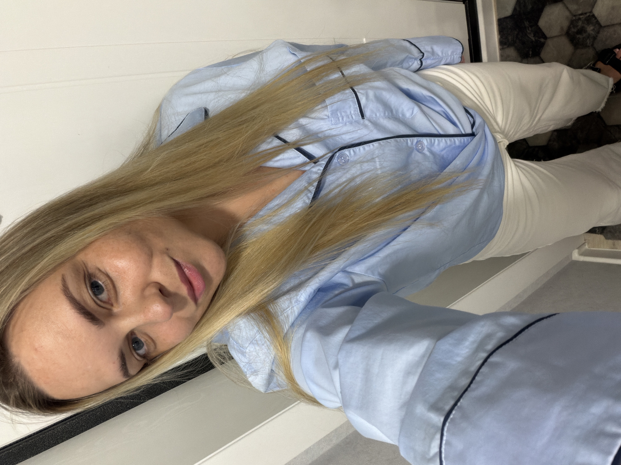A blonde woman, appearing to be in Votkinsk, Russia, is captured in a selfie taken indoors. She has long, straight, light blonde hair that cascades down her front, blue eyes, and fair skin. Her expression is neutral to slightly smiling, with her lips softly parted. She is wearing a light blue button-up shirt with dark piping details on the collar, placket, and cuffs, resembling a pajama top. She is also dressed in white or off-white trousers.
The background on the left consists of a plain white wall or door. On the right and towards the bottom of the image, a dark and light hexagonal tiled floor is visible, suggesting an interior space such as a bathroom or hallway. A dark frame or edge is also noticeable near her head and in the bottom right corner. The lighting is ambient and characteristic of an indoor setting, not indicating a specific time of day. No explicit objects other than her clothing are discernible, and there is no visible text within the image. The overall scene portrays a casual and relaxed moment.