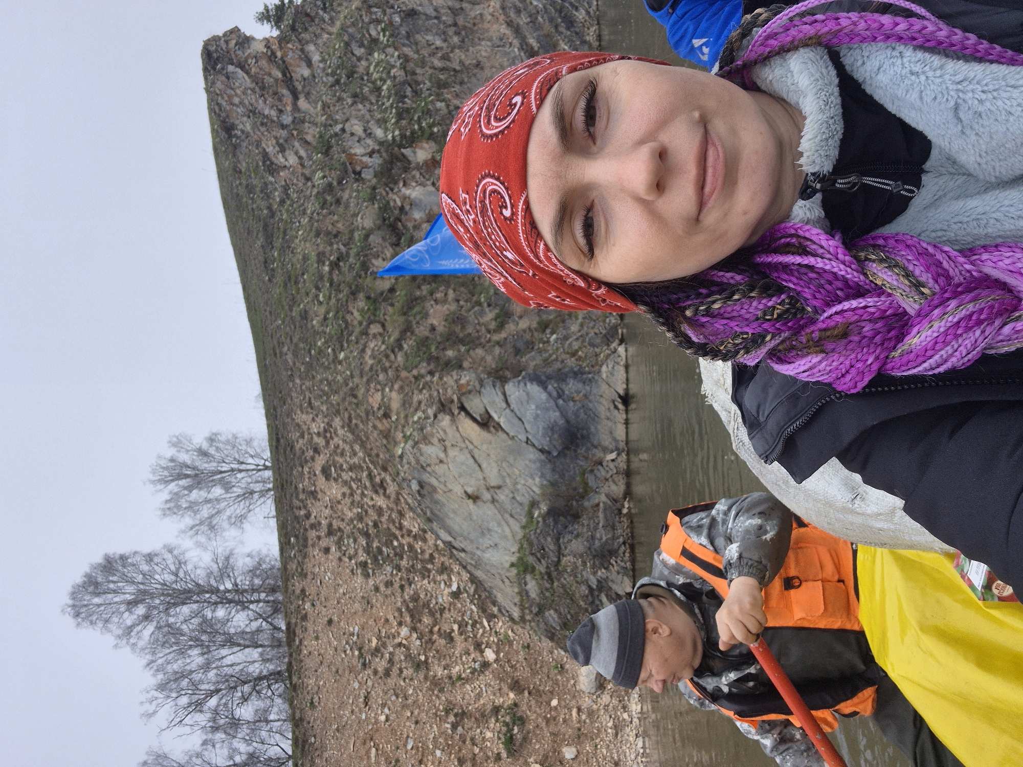 This image, rotated 90 degrees counter-clockwise from its original orientation, depicts two people engaged in a water activity in Новомунасипово, Russia, against a backdrop of a rocky landscape and overcast sky.
In the foreground, a woman looks directly at the camera with a gentle smile. She wears a red bandana with white paisley patterns, a light blue fuzzy jacket over a dark layer, and has long, prominent purple and dark braided hair. A small blue flag or piece of fabric is visible behind her head.
To her right, a man is partially visible, oriented away from the camera, actively paddling a yellow inflatable boat or raft with a red paddle. He is dressed in a gray and black striped beanie, a camouflage-patterned jacket, and a bright orange life vest. Next to him, a small package is visible with the text "GRAPE JUICE" and "100%" printed on it.
The setting includes a calm body of water, likely a river or lake, reflecting the grey, overcast sky. A steep, rocky bank or cliff, sparsely covered with vegetation, rises from the water's edge. Further in the background, several bare-branched trees stand against the pale sky. The attire of the individuals, including jackets and a beanie, suggests cool weather, and the bare trees indicate a non-growing season, such as late autumn, winter, or early spring. The scene captures an outdoor recreational excursion.