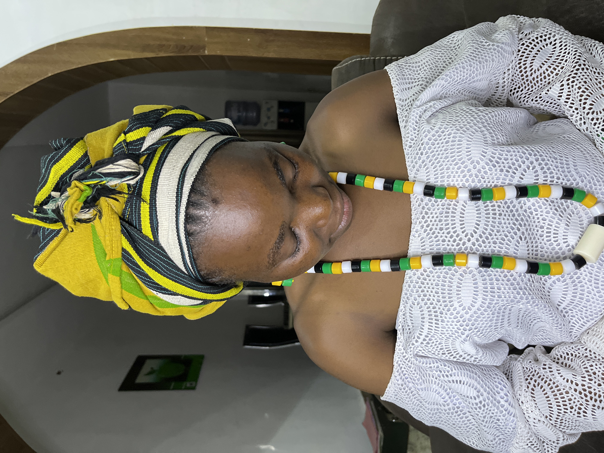 The image shows a woman with dark skin, wearing a colorful headwrap of yellow, green, black, and white stripes. She is also wearing a necklace with alternating black, green, white, and yellow beads. She is wearing a white, lacy garment. The setting appears to be indoors, with a glimpse of a dark wooden archway and possibly a television in the background. The location given is Jos, Nigeria.