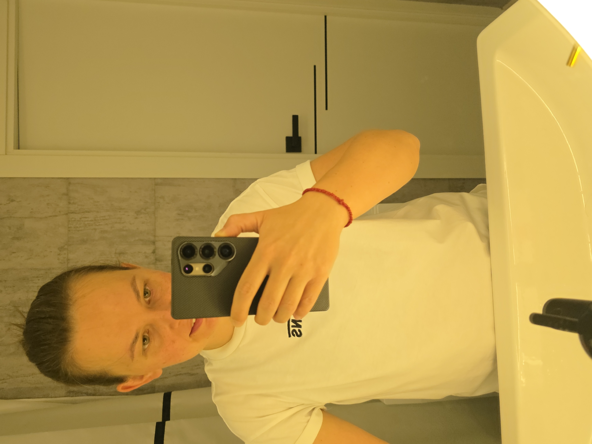 The image shows a person taking a selfie in a bathroom. The person is holding a smartphone in their right hand, with the phone's camera visible. They are wearing a white t-shirt with a black logo. A red beaded bracelet is visible on their wrist. The bathroom features tiled walls and a white sink in the foreground. A door with a black handle is visible in the background. The lighting is warm and yellow, casting a glow on the scene. There is no indication that this image was taken in Rostov-on-Don, Russia.