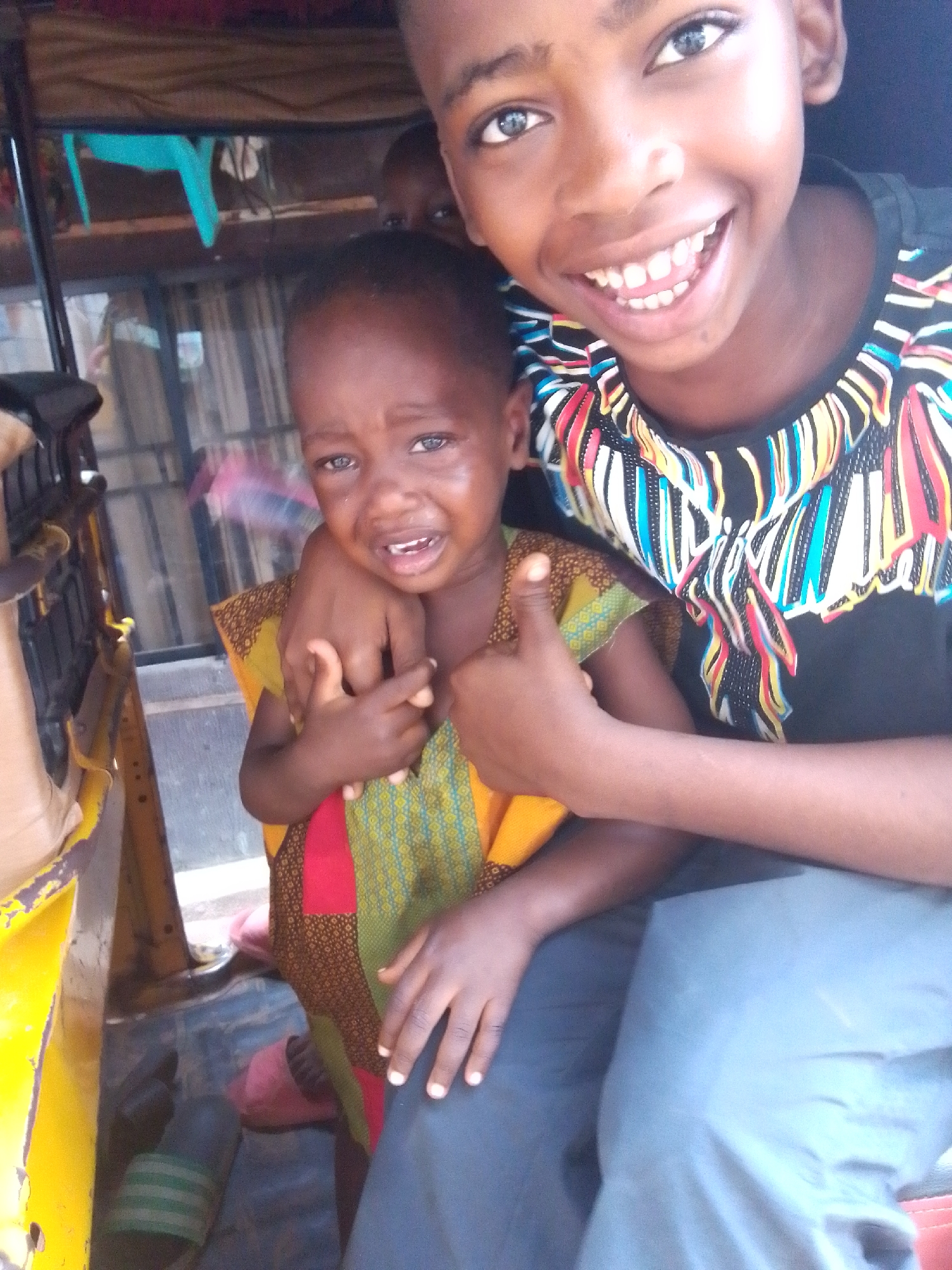 The image depicts two children inside what appears to be a three-wheeled vehicle, potentially an auto-rickshaw or "keke", common in Nkpor, Nigeria. One child, older, is smiling broadly at the camera. He is wearing a black T-shirt with multicolored embellishments. The other child, younger, is crying and looking at the camera. The younger child wears a colorful traditional garment. The background shows the interior of the vehicle and hints of an urban environment outside. One sandal is visible on the floor of the vehicle.