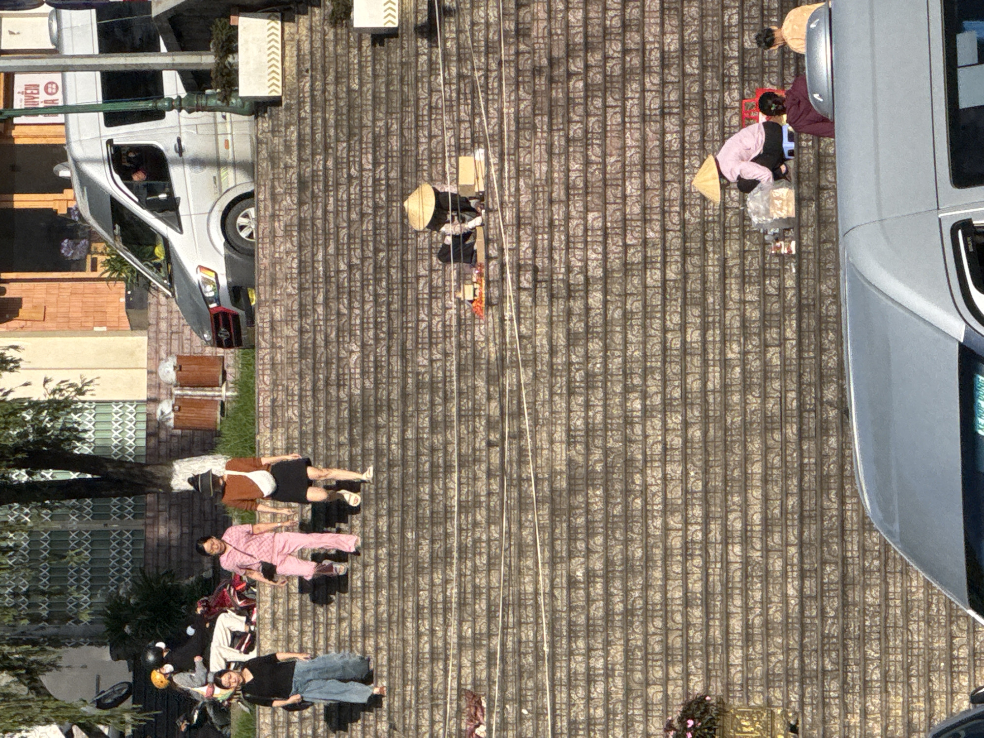 The image shows a wide stone staircase in Da Lat, Vietnam, with several people present. The stairs are made of textured stone tiles.
In the upper left, a motorcycle is parked, with two people on it wearing helmets. To their right, three women are walking down the stairs. The woman on the far left wears a black top and blue pants. The middle woman wears a pink patterned outfit. The woman on the far right wears a brown top, a dark skirt, and a hat, with a white bag slung across her chest.
Further down the stairs, two individuals are sitting. One person, wearing a conical hat, is surrounded by various items, possibly for sale, including boxes and small red containers. Another person, also wearing a conical hat, is seated on a small blue stool and appears to be interacting with some goods. To their right, two other individuals are visible, one wearing a burgundy hooded jacket and the other a yellow top.
In the background, a silver van is parked, and parts of buildings and trees are visible. The lighting suggests it is daytime, possibly late afternoon, with strong shadows cast on the stairs. The overall scene depicts everyday life and commerce on a public staircase in Da Lat.