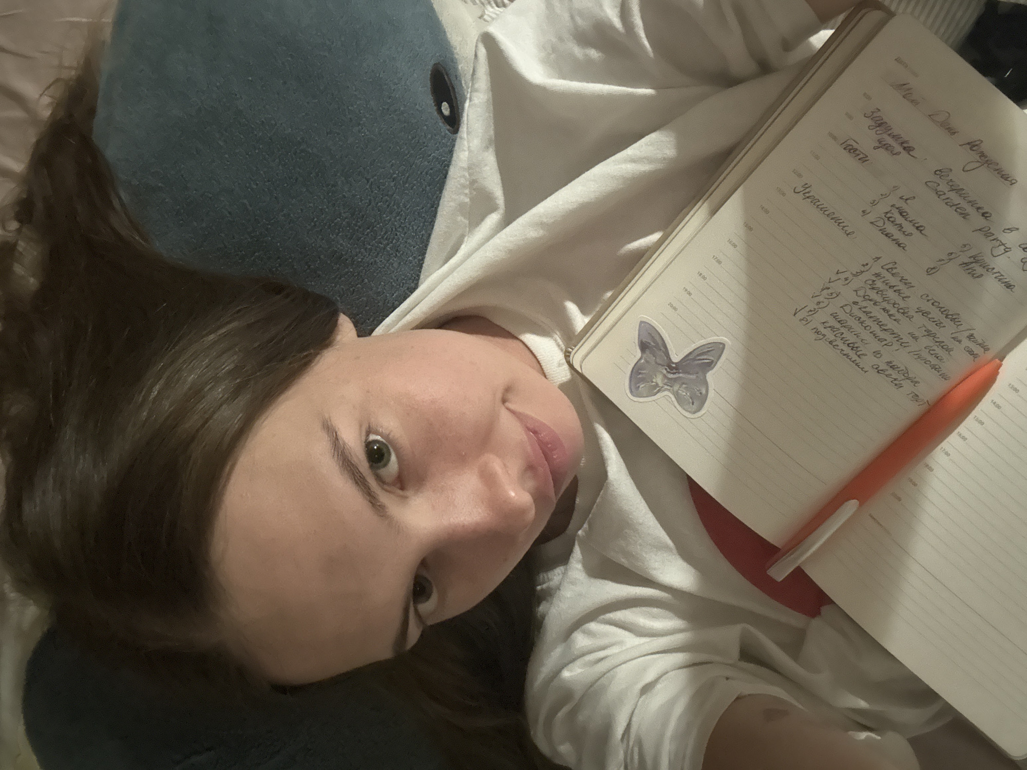 A young woman with brown hair lies on her back, holding an open notebook and an orange pen. She is wearing a white t-shirt. The notebook contains handwritten text and a sticker of a purple butterfly. The background consists of a dark blue pillow. The image is taken from a close-up perspective, focusing on the woman's face and the notebook. There is no discernible urban environment or location within the city of Perm, Russia, visible in the image.