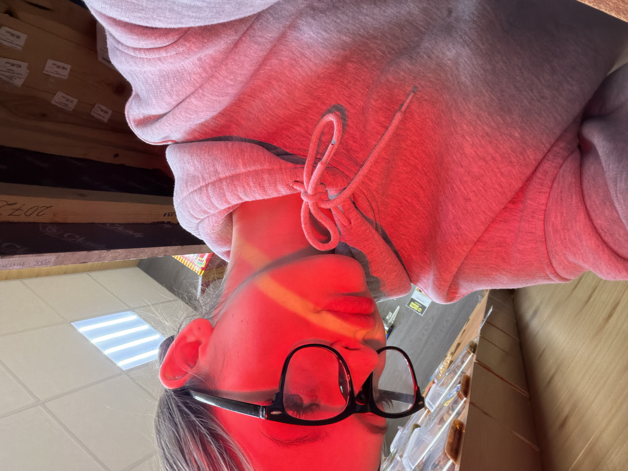 The image captures a close-up, low-angle perspective of a person indoors in Semyonov, Russia, looking upwards, dramatically illuminated by a strong red light. The person, likely young, is wearing a light-colored hooded sweatshirt with visible drawstrings tied in a bow, and black-framed glasses. Their face is bathed in a vivid red glow, with a distinct horizontal band of brighter light cutting across their forehead and eyes, suggesting an external light source or reflection.
The unusual camera angle presents the ceiling and surroundings as if viewed from below. A tiled ceiling with rectangular fluorescent light panels is visible above the person's head. To their left (when oriented upright), there is a wooden structure, possibly shelving or beams, featuring several small white labels. One label clearly displays "148.00," likely indicating a price or inventory number. Other labels appear similar. Below this, a wooden beam has "2072" and partially "#EV" inscribed or printed on it. To the person's right, more wooden surfaces, potentially shelves or walls, are present. A partially visible yellow label with black text, possibly "SMT" or "BMT", can be seen.
The activity depicted is simply the person looking up. The red lighting is the most notable detail, giving the scene a distinct, perhaps artificial, ambiance. The location cues, such as the fluorescent lights and labelled wooden structures, suggest a commercial environment like a store or warehouse. No specific time of day or emotional state is discernible.