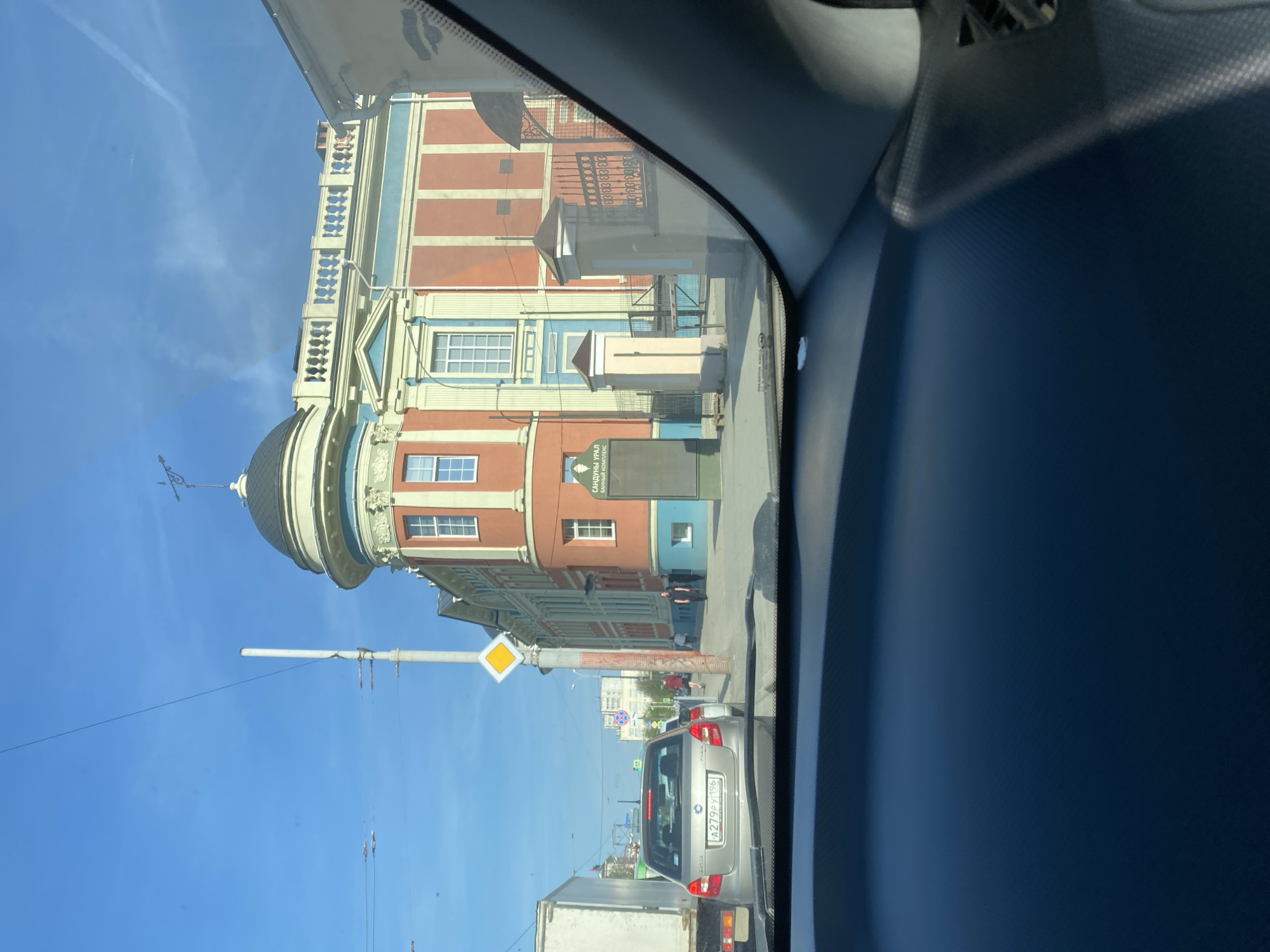 The image captures a view from inside a vehicle, looking out towards a street in Yekaterinburg, Russia. A prominent building with a dome and a multi-colored facade of orange, blue, and cream dominates the view. The building is identified as "Sanduny Ural," which is a bathhouse. A yellow diamond-shaped street sign is visible near the building. Below, traffic flows, including a silver car with license plate "A279PM196" and a white truck. Pedestrians are walking along the sidewalk in front of the building. The sky is blue with some wispy clouds. The interior of the vehicle, including the dashboard, is partially visible on the right side of the frame.