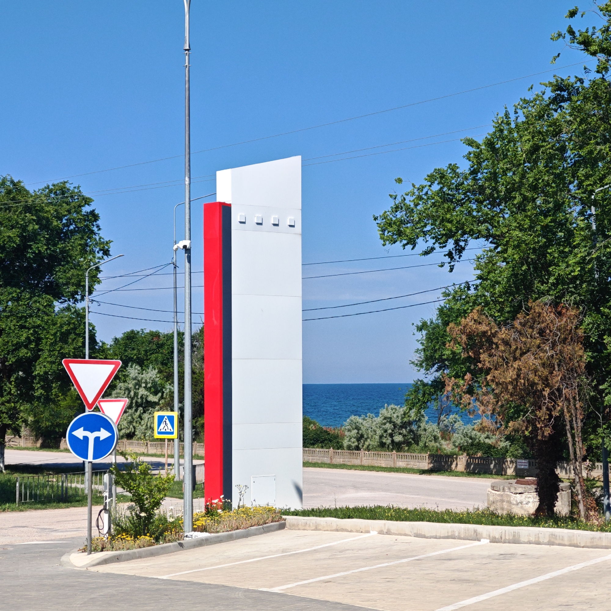 In the city of Pervomaiske settlement, Ukraine, on a clear, sunny day, a modern, tall, rectangular structure with a red vertical stripe on its left side stands prominently. This structure is flanked by a metal pole with a security camera and a street lamp. To the left of the structure, a series of traffic signs are visible: a large, white, inverted red triangle indicating yield, a smaller similar sign below it, and a blue circular sign with two opposing arrows, signifying a choice between turning left or right. A yellow square sign with a pedestrian symbol is also present. In the foreground, a paved parking lot with white lines is delineated by a concrete curb. Beyond the parking lot, a road stretches into the background, separated by a low metal fence and a line of small green and yellow flowering plants. Lush green trees border the scene on the left and right, with a glimpse of the blue sea visible in the distance between the trees and the road. The environment suggests a public space, possibly near a coastal area, with a focus on traffic management and modern urban design.