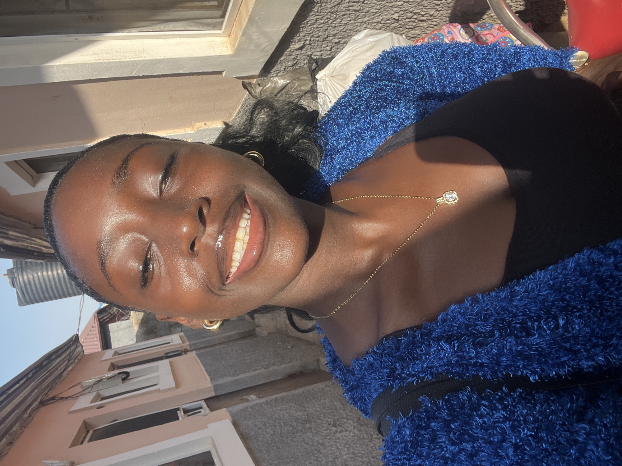 A medium-shot outdoor portrait shows a smiling dark-skinned woman wearing a blue fuzzy jacket and a black top, with a gold necklace and hoop earrings. She is on a street in Abuja, Nigeria. The background includes residential buildings, a metal structure, and a street with various objects and trash. Sunlight highlights her face and neck.