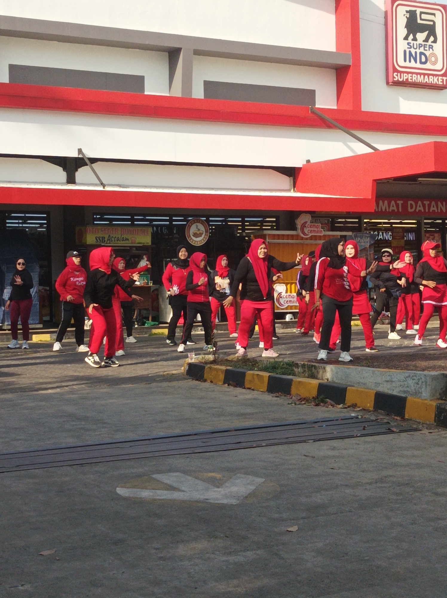The image captures an outdoor aerobics class in front of a Super Indo supermarket in South Jakarta, Indonesia. A group of approximately ten women, all dressed in athletic wear consisting of red tops and pants or skirts, and most wearing red hijabs, are engaged in synchronized exercises. Their movements are dynamic, with arms extended and legs bent, suggesting active participation in a choreographed routine. The supermarket facade is prominent in the background, featuring a red and white color scheme with the "SUPER INDO" logo and the word "SUPERMARKET" clearly visible. A partial view of a sign above the entrance reads "AMAT DATANG," which is likely part of a welcome message in Indonesian. The ground is paved asphalt with yellow and black striped concrete barriers bordering the exercise area. The lighting suggests it is daytime, with shadows indicating the sun's position. The overall scene depicts a community fitness activity taking place in a semi-public commercial space.
