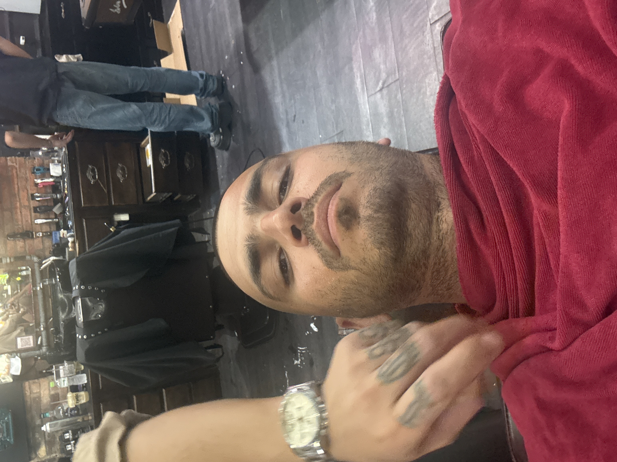 The image presents a man undergoing a grooming service, possibly a shave or facial treatment, in what appears to be a barbershop or salon setting. He is partially covered with a red towel. A person with tattoos on their hand is attending to him, possibly applying a product or wiping his face. The attendant is wearing a watch. The background reveals elements of a barbershop, including shelves with grooming products, equipment, and a mirror. Another person is seen in the background wearing jeans and a t-shirt. The location cannot be determined from the image. Đà Nẵng, Vietnam cannot be confirmed as the setting.