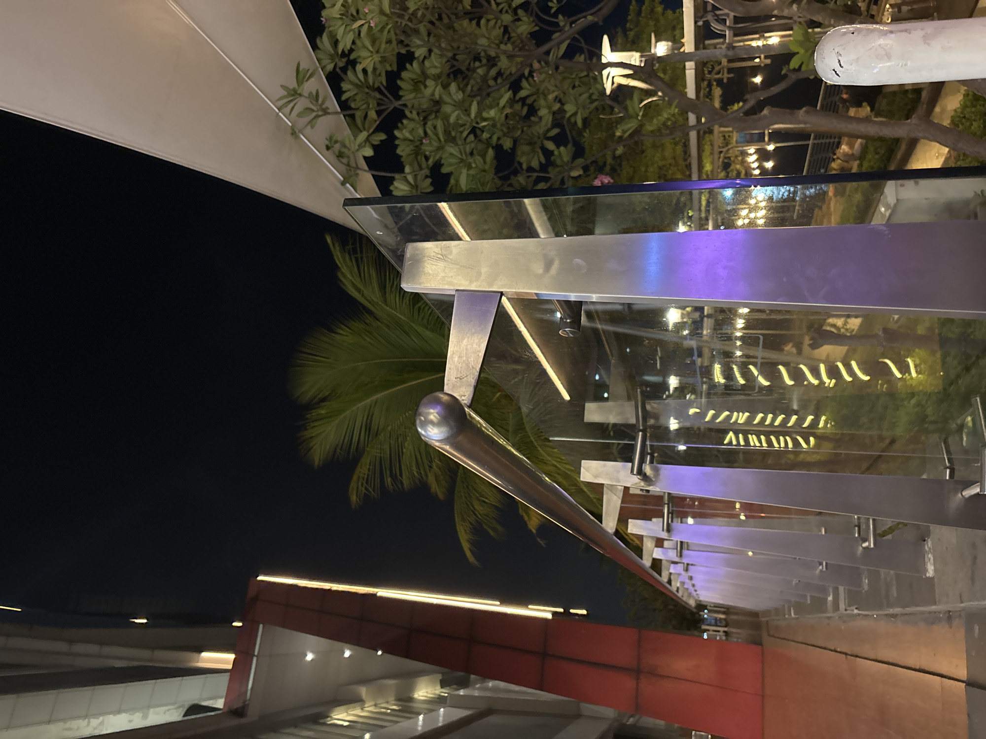 The image depicts an outdoor walkway or balcony at night, likely in North Jakarta, Indonesia, given the prominent presence of palm trees and tropical foliage. On the left, a modern building with red and silver accents and strip lighting is visible, suggesting a commercial or residential complex. A metal railing with glass panels runs along the walkway, with purple lighting emanating from some of the supports. Beyond the railing, a body of water, possibly a canal or bay, can be seen with lights reflecting on its surface, indicative of a waterfront location. Trees with lush green leaves and some small pink flowers are present, adding to the tropical ambiance. The sky is dark and clear, typical of a nighttime scene. The overall impression is a well-lit, modern urban environment with a touch of nature.