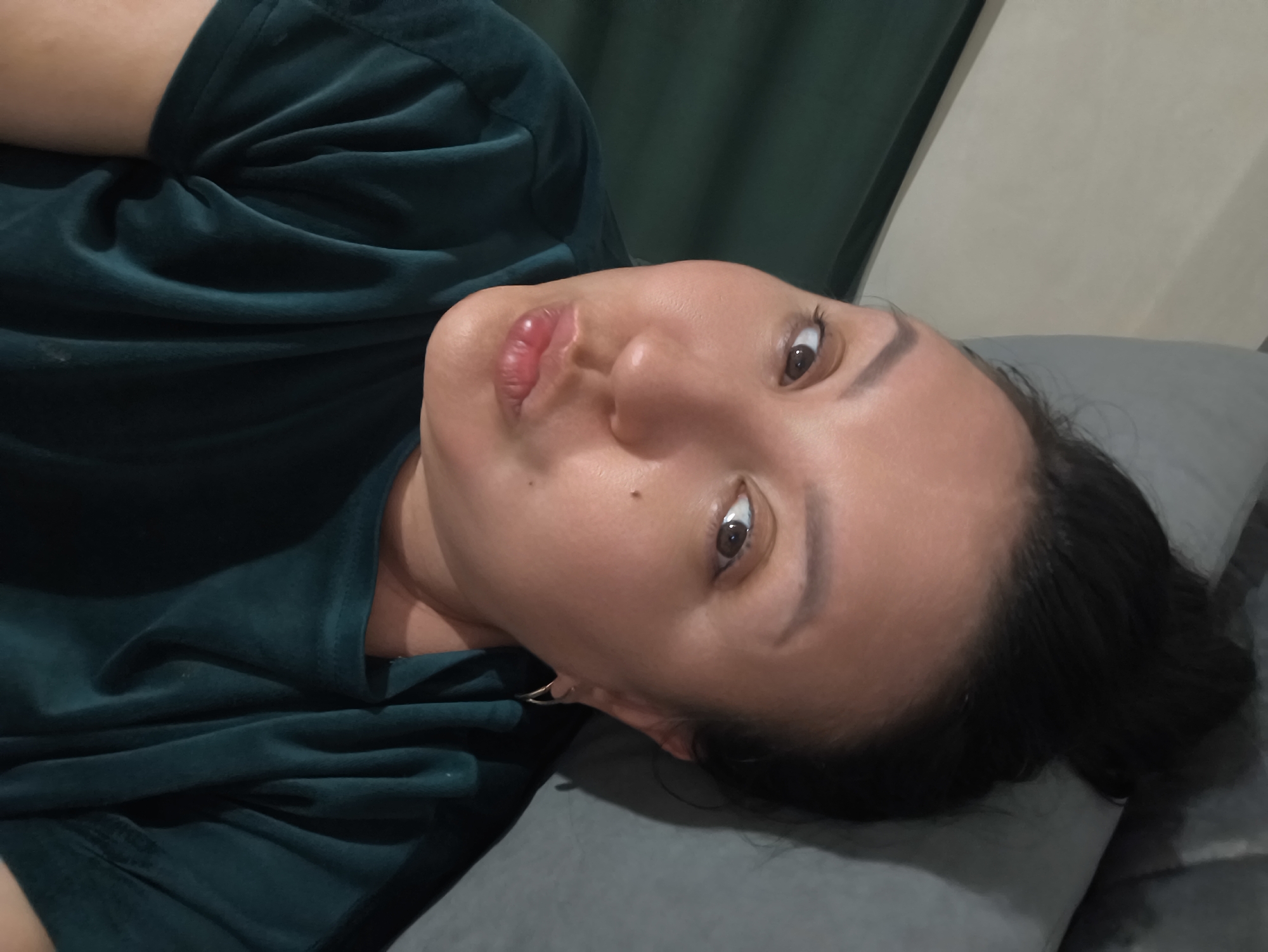 A woman is lying on a grey pillow, wearing a dark green velvet shirt. She is looking at the camera. The background includes a green curtain and a beige wall. The picture provides no indication that it was taken in Сокуры, Russia.