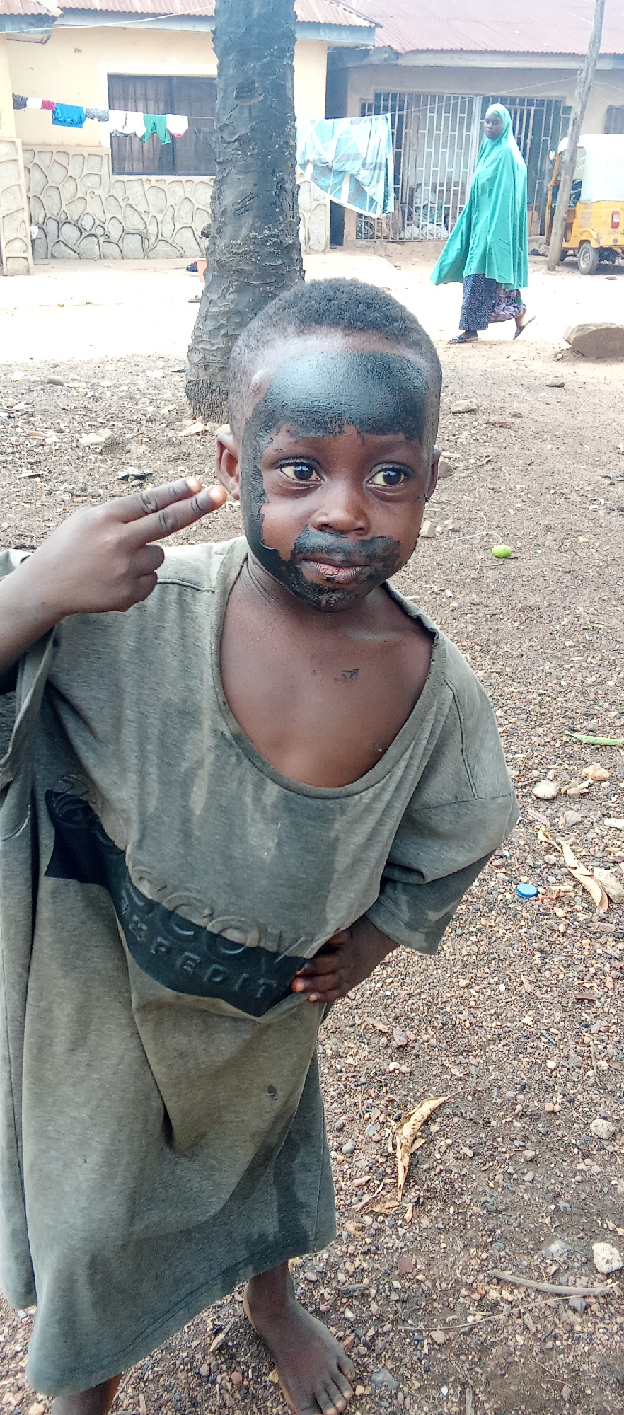 A young child stands outdoors in Akwanga, Nigeria, with black markings covering their forehead and resembling a beard. The child wears a large, soiled gray t-shirt. They are making a gesture with their hand, extending two fingers as if holding a gun. The ground is covered with small rocks and debris. In the background, there is a building with laundry hanging on a line, a woman wearing a green hijab and a yellow tricycle. A palm tree is next to the child.