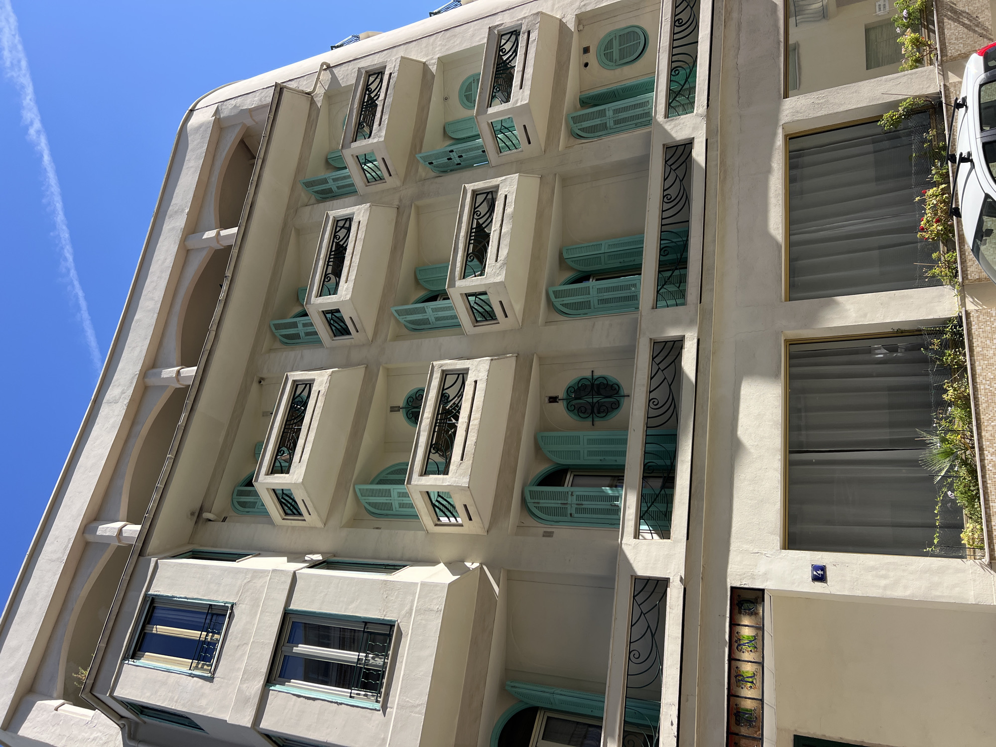 The image shows a multi-story building in Nice, France, with a bright blue sky overhead. The building has a light-colored facade and features multiple balconies with light turquoise shutters. The balconies have decorative black metal railings. There are several windows on the building, some with closed shutters and others with open shutters. The ground level of the building has large windows with sheer curtains. A white car with a roof rack is parked in front of the building, partially obscuring the lower windows. There is greenery and potted plants visible on the balconies and in front of the ground-level windows. A blue number "4" is visible on the facade above one of the ground-level windows. The building appears to be an apartment building or a hotel.