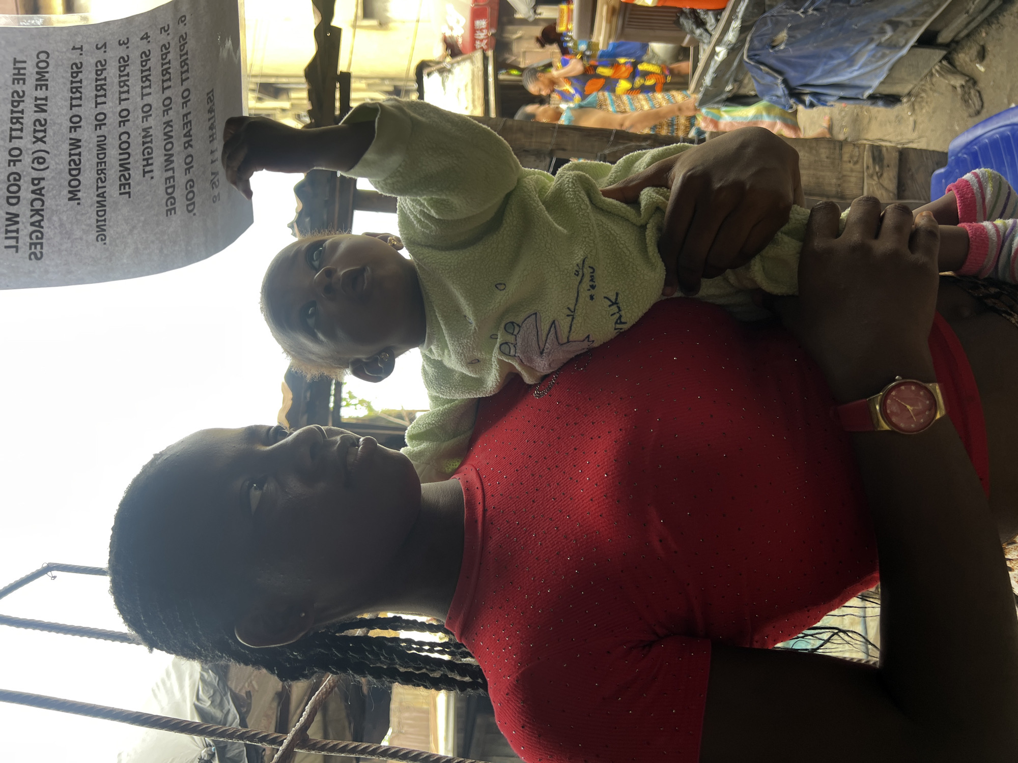 A young Black woman stands outdoors, holding a baby. The woman wears a red, sparkling t-shirt and a watch with a red band and gold face. The baby, dressed in a pale green outfit with a frog graphic and striped socks, reaches up towards a piece of paper with text. The text, written in a reversed font, appears to be a religious passage, potentially from Isaiah. The background shows a bustling market scene in Nkpor, Nigeria, with other people, stalls, and corrugated metal structures. The lighting suggests it is daytime.