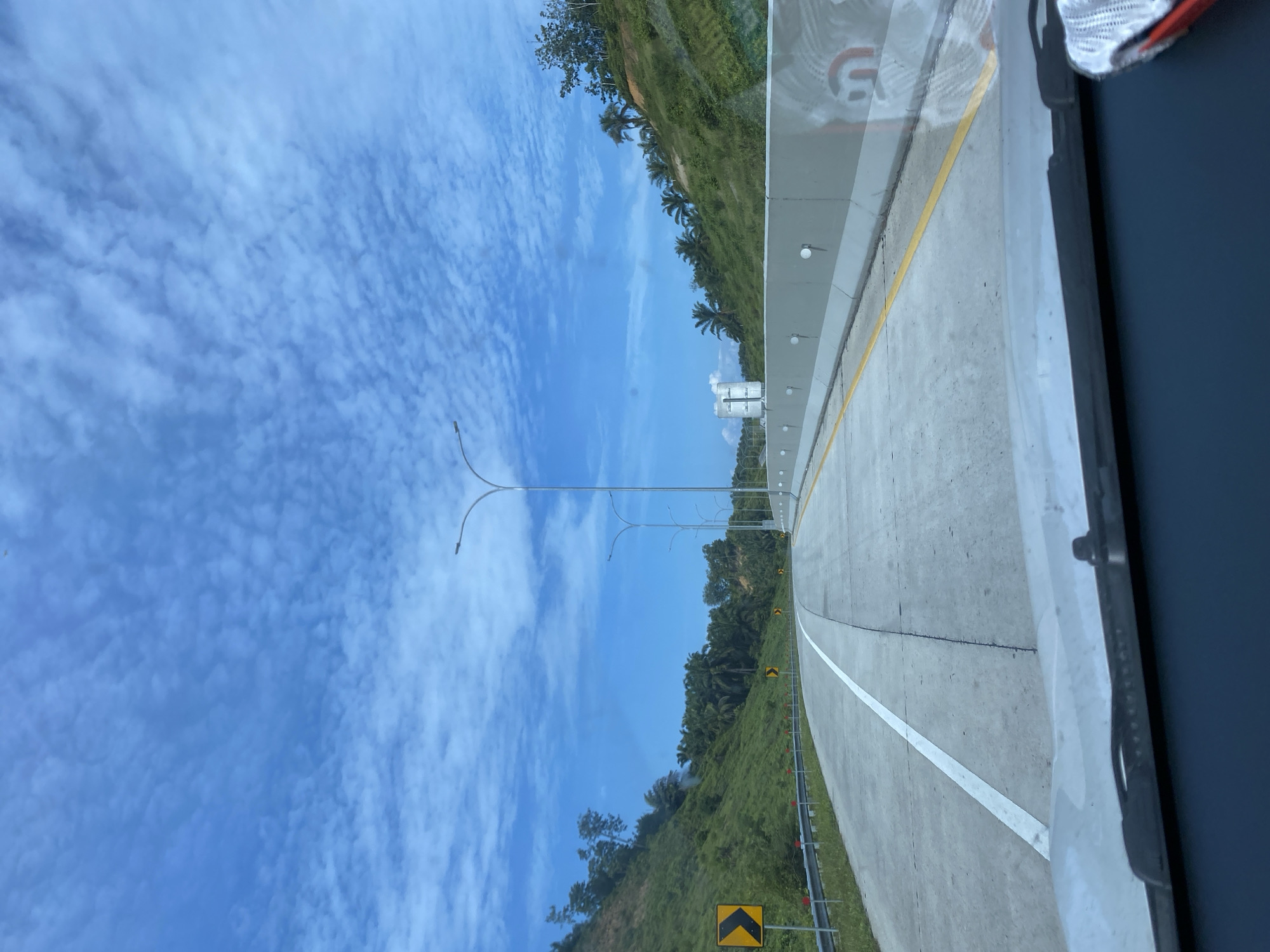 The image shows a view from inside a vehicle traveling on a highway. The road is paved and has clear white lane markings. On the left side of the road, there is a grassy embankment with trees and vegetation. A yellow sign indicating a curve is visible on the left. On the right side of the road, there is a concrete barrier. The sky is blue with scattered white clouds. Palm trees and other tropical vegetation are visible on the hillsides. Streetlights are spaced along the right side of the highway. The scene appears to be in a rural or semi-rural area. Based on the provided information, this scene is located in the city of Tangkahan Durian, Indonesia.