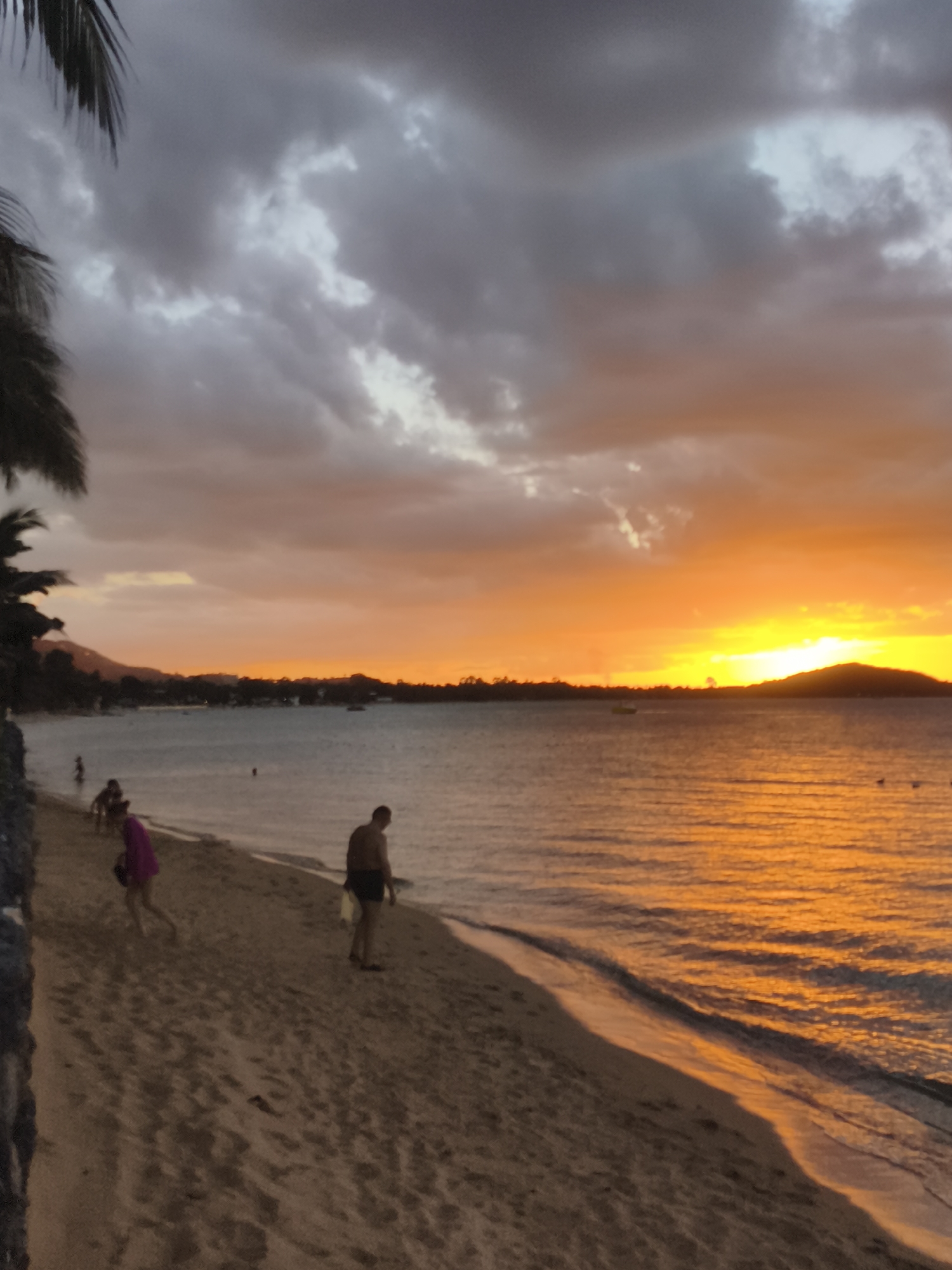The image depicts a tropical beach scene at sunset in Baan Bo Phut, Thailand. The sky is filled with dramatic clouds in shades of grey, pink, and orange, illuminated by the bright yellow and orange hues of the setting sun. The sun is low on the horizon, casting a golden reflection on the calm water.
On the sandy beach in the foreground, several people are present. To the left, a person wearing a bright pink garment walks away from the camera. Further down the beach, a man in dark shorts stands facing the water. The beach itself shows faint footprints in the sand, and the edge of the water laps gently onto the shore. In the distance, across the water, a silhouetted shoreline with vegetation and buildings can be seen, and a few small boats are visible on the sea. Palm fronds are visible in the upper left corner of the image, framing the scene. The overall atmosphere is serene and tranquil.