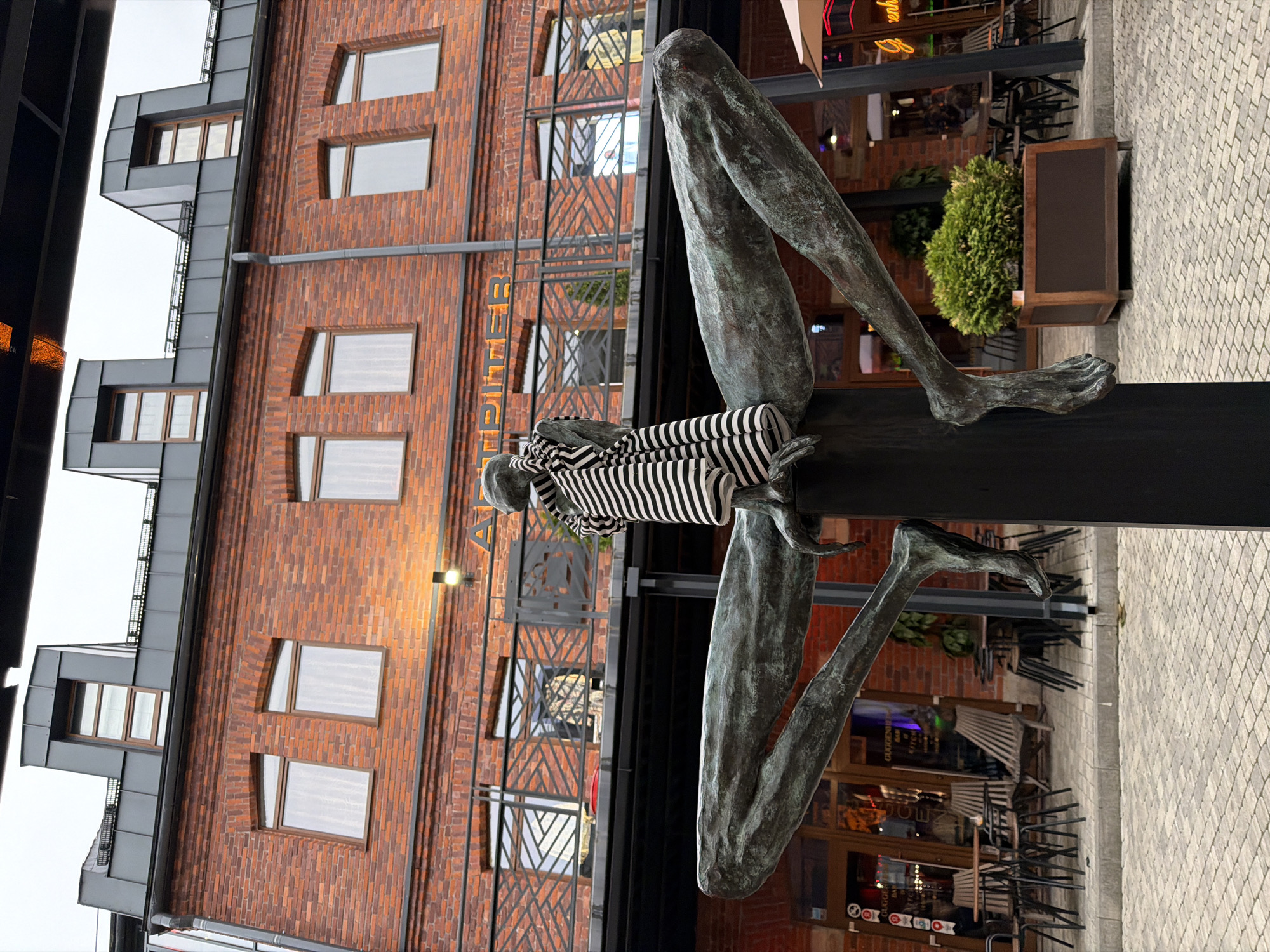 The image depicts a bronze statue of a seated figure with elongated limbs, wrapped in a striped cloth, located in an urban area. The background shows a brick building with multiple windows, typical of historical architecture. Tables and chairs are visible, suggesting an outdoor seating area for a cafe or restaurant. The location is identified as Saint Petersburg, Russia. The statue appears to be a contemporary art installation.