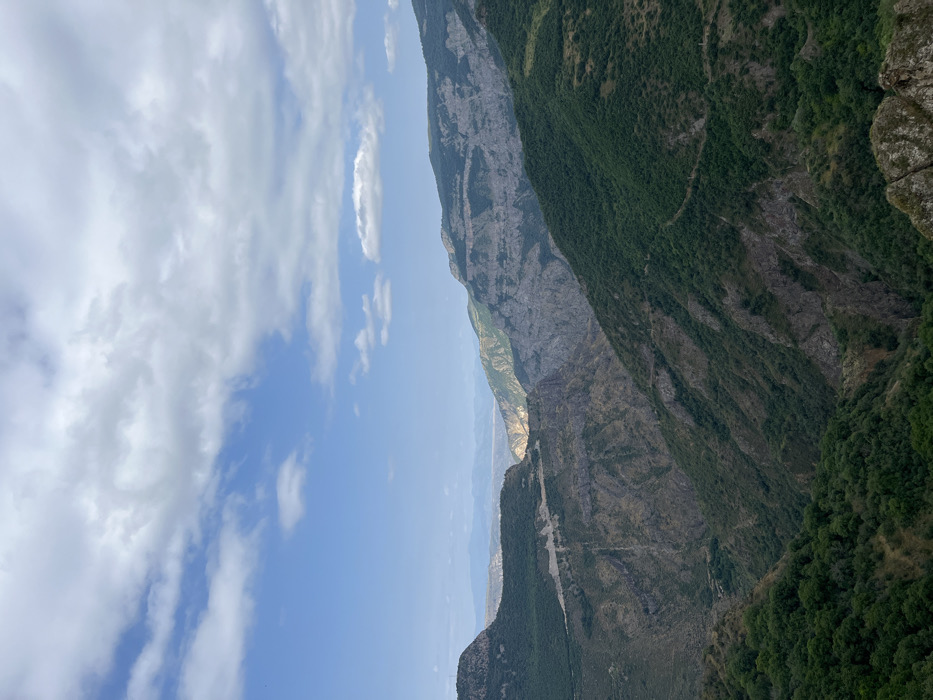 The image displays a panoramic view of the Vorotan River Gorge in Tatev, Syunik Province, Armenia. The scene is dominated by a deep, rugged gorge featuring steep mountain slopes. These slopes are heavily covered in dark green vegetation, interspersed with visible sections of grey and brown rock. A light-colored, winding road or track is discernible traversing a forested slope on the left side of the gorge. In the background, distant mountains appear hazy, with a distinct sunlit area illuminating a segment of the valley floor. The sky above is blue with scattered white cumulus clouds. Dark, angular rock outcrops are present in the immediate foreground on the lower right. No human or animal figures are visible within the frame.