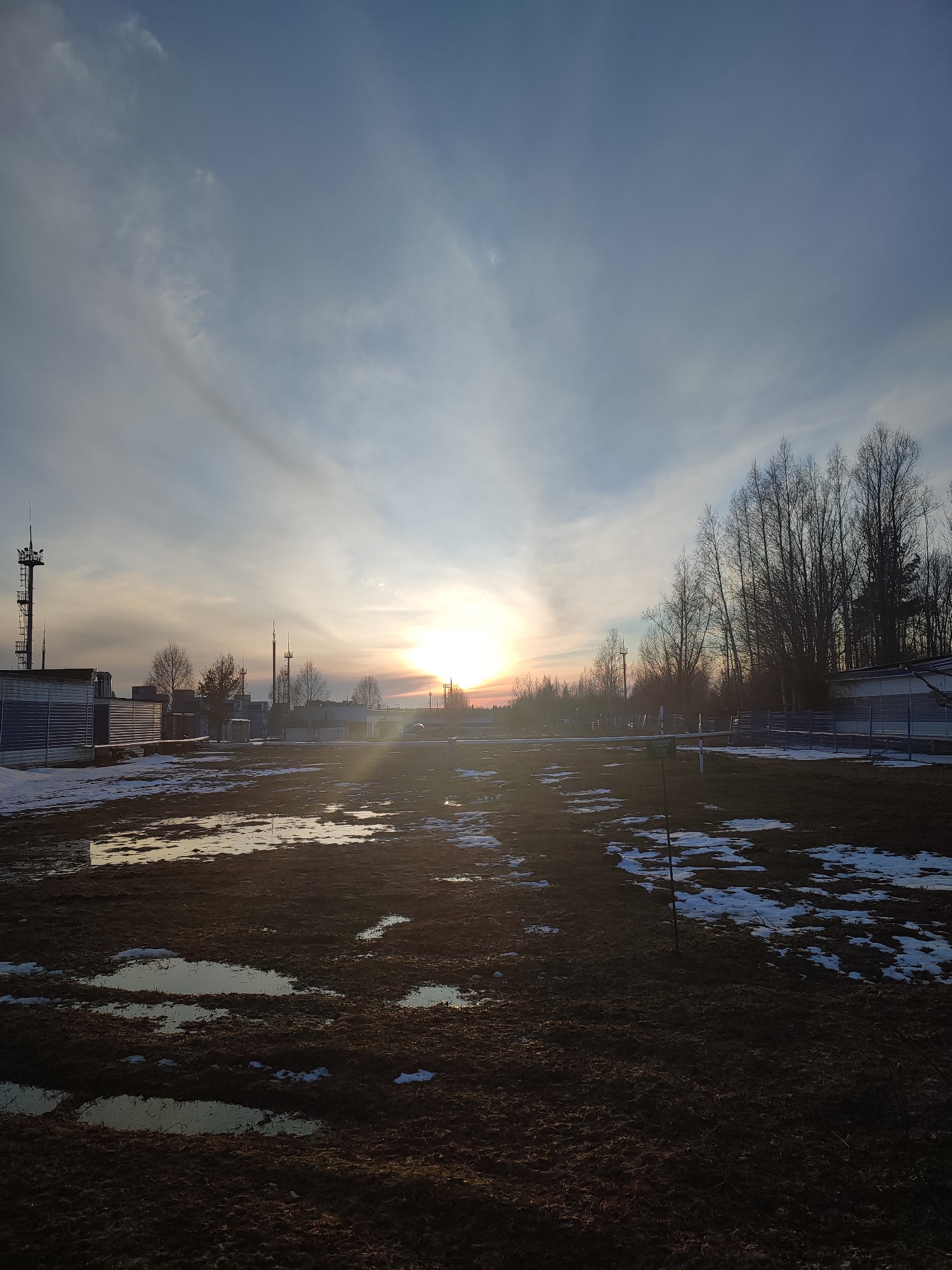 The image depicts a landscape at sunset. The ground is a mix of dark soil, melting snow patches, and standing water in puddles. In the middle ground, there is a line of trees and several buildings, some appearing to be industrial or storage structures. Two communication towers stand prominently on the left side. The sky is a gradient of blues and yellows with the sun setting on the horizon, casting a bright glow. The location is a city in Unknown, Unknown.