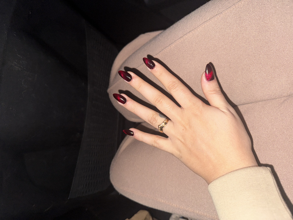 A woman's hand rests on a pair of tan-colored pants, possibly in a car. Her nails are almond-shaped with a dark, red-to-black ombre design, giving a striking appearance. A gold-colored ring adorns her ring finger. The background is partially shadowed, with a hint of the car's interior visible. The sleeve of her beige-toned top is also visible.