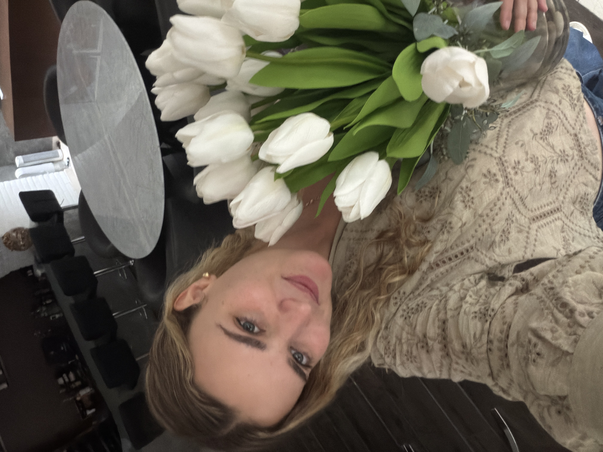 A woman with blonde wavy hair and blue eyes is holding a vase of white tulips. She is wearing a beige patterned blouse. The background includes a round glass table, a black chair, and a dark wood floor. In the distance, there is a rack with various items. The location is not San Bernardino Tlaxcalancingo, Mexico.