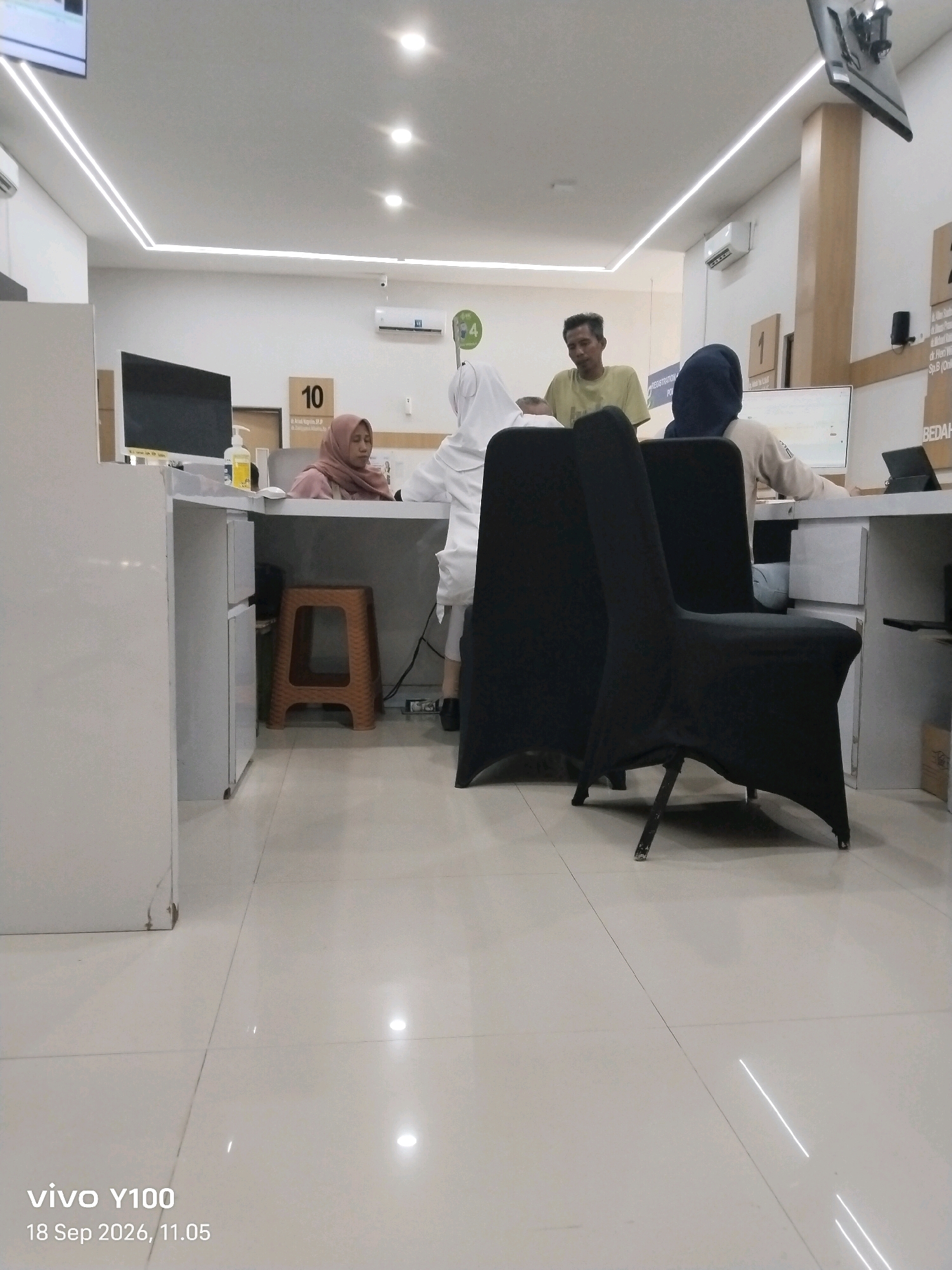 In a bright, modern interior, possibly a reception area or service counter, located in Situbondo, Indonesia, three individuals are visible. A woman wearing a pink hijab sits at a counter, looking down. In front of her is a bottle of hand sanitizer. To her right, another woman wearing a dark blue hijab is seated at a computer workstation, her back to the camera. A man in a light yellow t-shirt stands behind the counter, looking towards the seated women. Two chairs, covered in black fabric, are positioned in the foreground. The environment features white tiled flooring and recessed ceiling lighting. A digital display is mounted on the wall in the background, and a sign with the number "10" is visible above the counter. The timestamp at the bottom of the image indicates "vivo Y100 18 Sep 2026, 11.05."