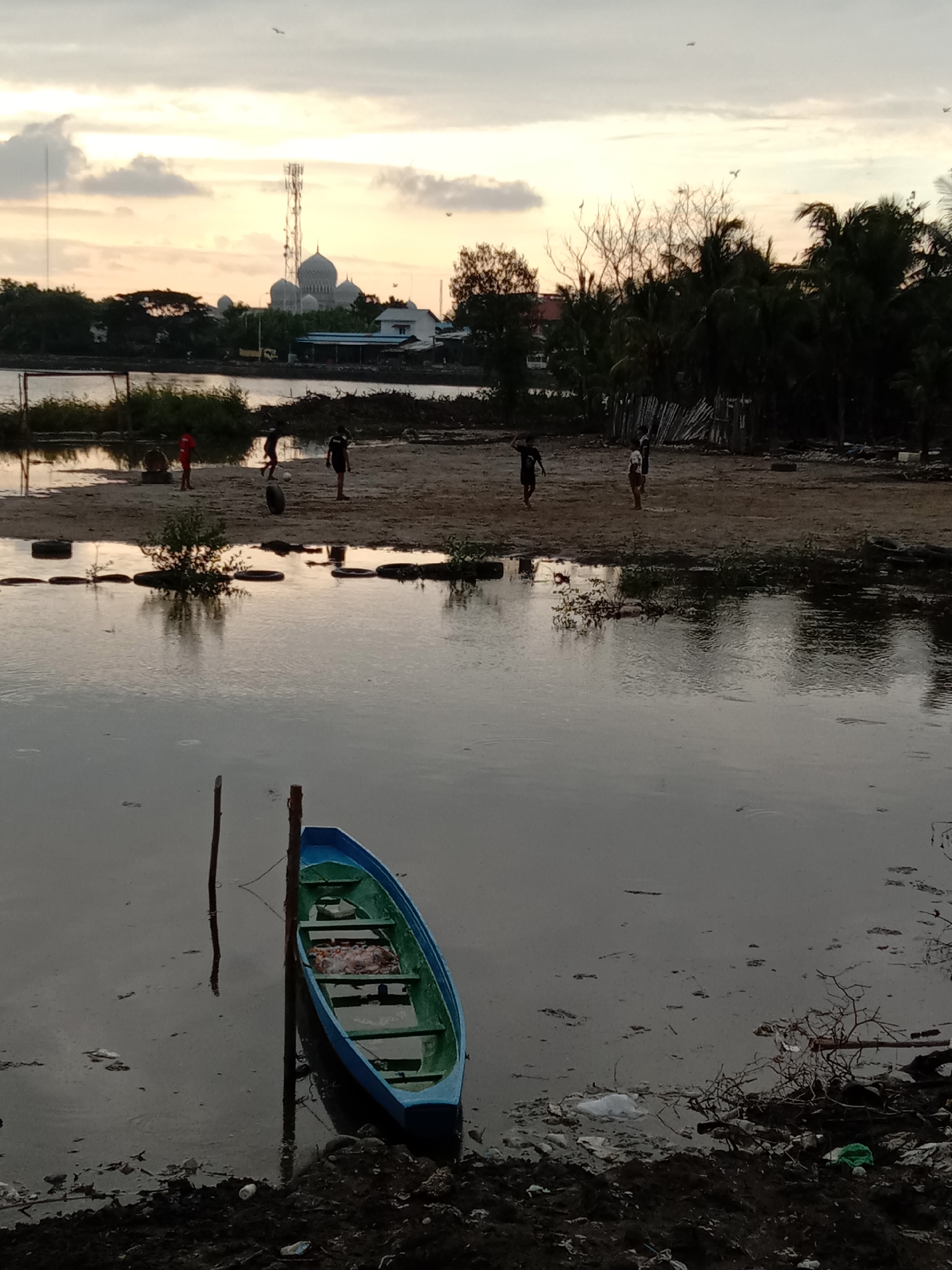 This image depicts a coastal or waterfront scene at dusk in Lhokseumawe, Indonesia. The sky above is a gradient of pale orange and grey, with scattered clouds and several birds in flight.
In the mid-distance, a modern mosque with multiple grey, ribbed domes and a prominent communication tower rise above the tree line. Various buildings and dense tropical foliage are visible along the distant shore.
The midground features a muddy, sandy strip of land separating two bodies of water. On this strip, approximately five children, all appearing to be male, are engaged in playing. One child in a red shirt stands near a makeshift soccer goalpost on the left. Other children in dark clothing are scattered across the field; one is actively kicking a small, light-colored ball, while another is positioned further right. Discarded tires and sparse vegetation are present on this land strip.
The foreground is dominated by a calm, greyish body of water that reflects the sky and the surrounding environment. A small, wooden boat, painted blue on the exterior and green on the interior, is moored to two dark wooden poles driven into the water on the left side. The boat appears to be a traditional local vessel, possibly for fishing or transport. The immediate shoreline in the foreground is muddy and strewn with considerable refuse, including plastic bags, bottles, and other debris, indicating an unkempt environment.