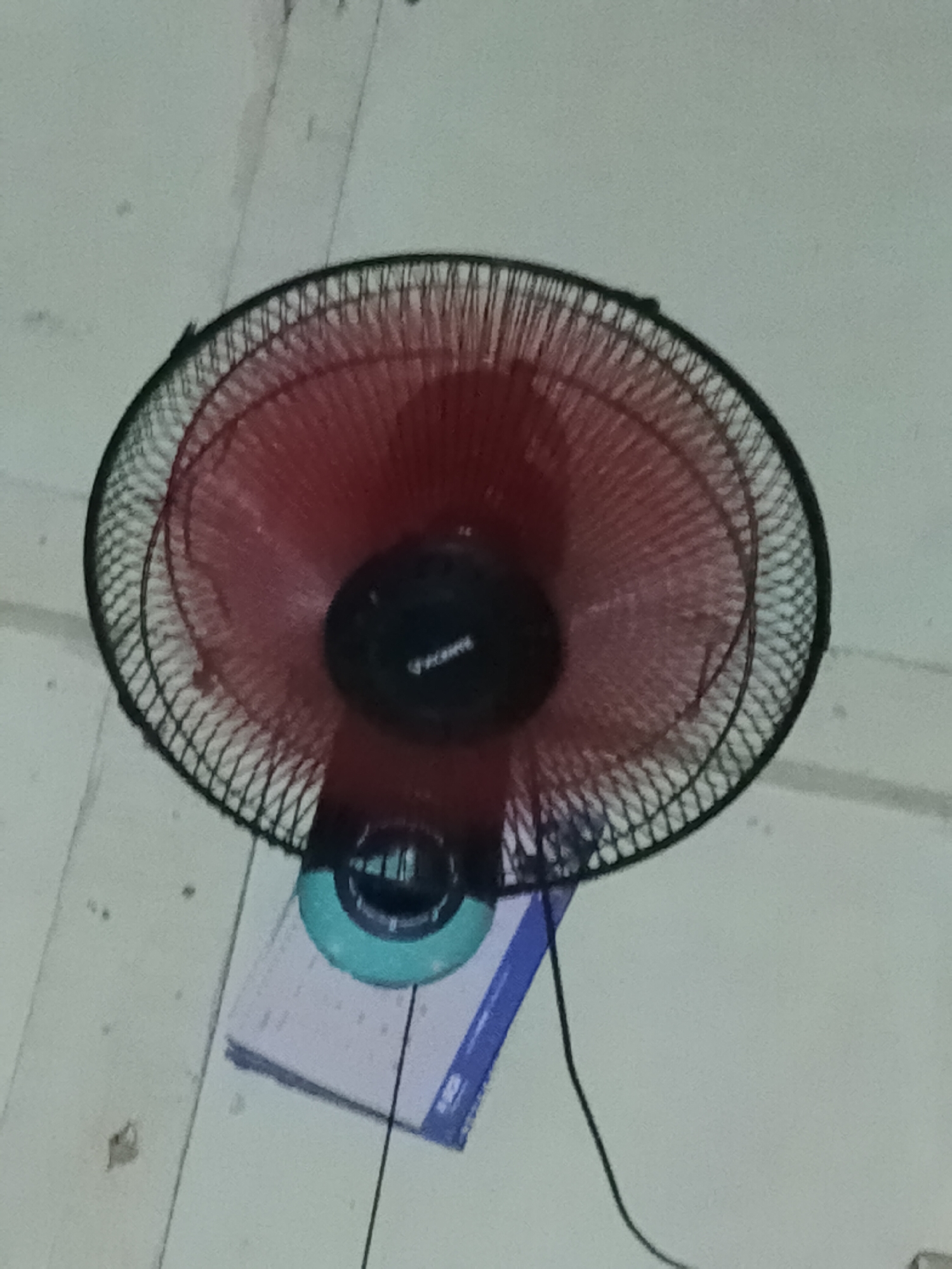An upside-down red fan is mounted on a white ceiling. The fan has a black grill and a black motor housing with a logo. Below the fan, a blue and white calendar is visible. The scene appears to be indoors, likely a room in a building in Bencongan, Indonesia. There are no people or animals visible in the image. The lighting is dim, and the image quality is low.