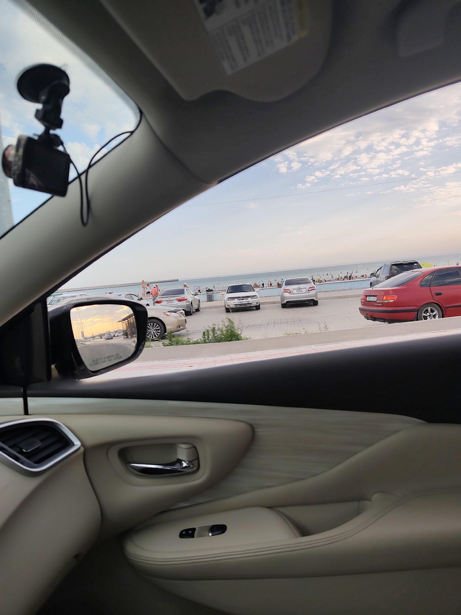 The image is a snapshot taken from inside a car, looking out towards a coastal area in Ömirzaq, Kazakhstan. The foreground shows the interior of the car, with a beige leather door panel, a chrome door handle, and a vent visible. A dashcam is mounted on the windshield.
Outside the car, the scene depicts a street bordering a beach. Several cars are parked along the street, and beyond them, people are gathered on the beach and in the water, suggesting a recreational setting. The sea stretches to the horizon under a cloudy sky. The side mirror of the car reflects a view of the street with traffic and people, illuminated by the warm glow of what appears to be sunset. The overall impression is a serene, everyday scene in a coastal town.