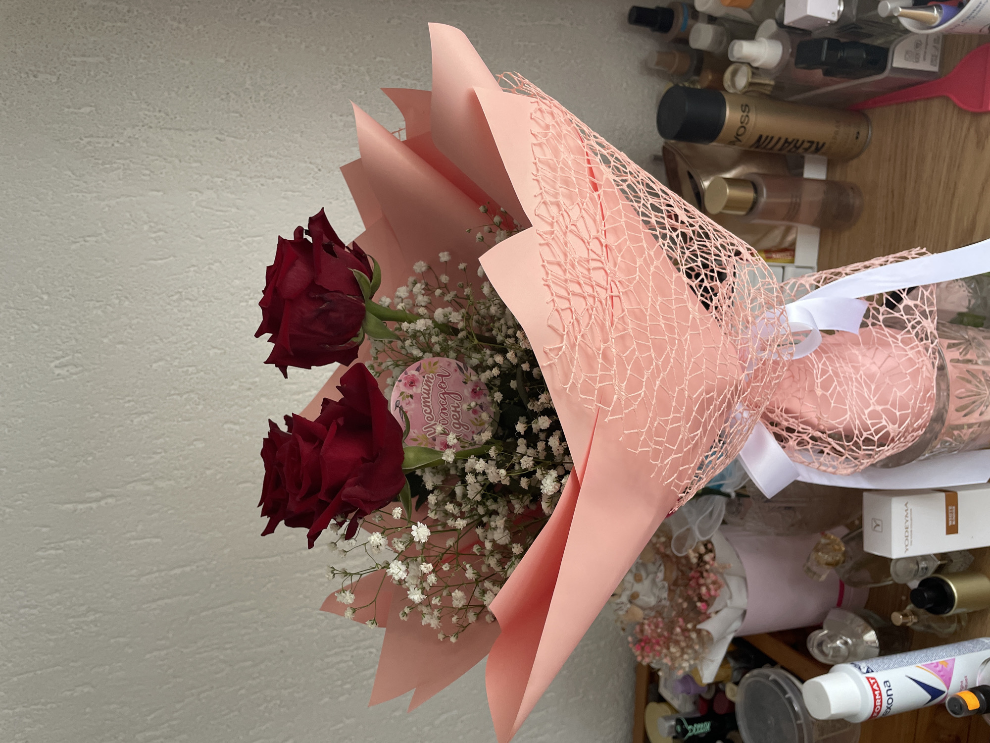 A bouquet of three red roses and baby's breath, wrapped in pink paper and tied with a white ribbon, sits on a surface cluttered with cosmetics and toiletries. The backdrop is a textured white wall. This image may have been taken indoors, potentially in Zlatograd, Bulgaria, although that cannot be confirmed definitively from the scene alone.