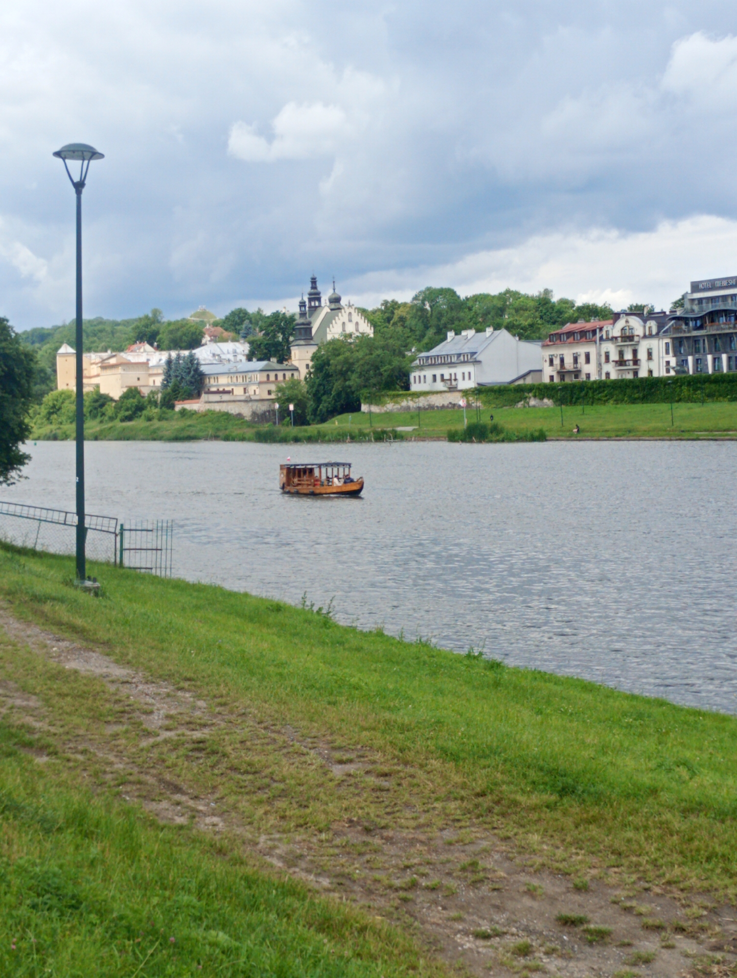 The image depicts a wide, calm river in Krakow, Poland. On the river, a small wooden tour boat with passengers is visible, moving to the right. The left bank of the river is lined with lush green grass and a dirt path, with a lamppost and a section of a fence also present. Across the river, a collection of buildings form the cityscape. These include what appears to be a historical complex with cream-colored walls and arched windows, possibly a castle or monastery, and other buildings with red and white facades. Further to the right, modern apartment buildings with balconies and a building with a sign that reads "HOTEL GROMADZKI" are visible. The sky is overcast with a mix of gray clouds and patches of lighter sky. The overall scene suggests a tranquil day in a city with a rich historical and modern architectural landscape along a prominent river.