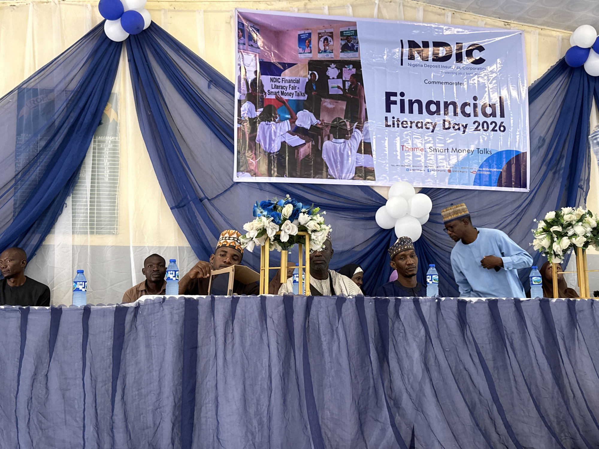 This media file depicts an indoor event, likely a financial literacy seminar or workshop, taking place in Unguwan Dosa, Nigeria. The scene is dominated by a long table draped in blue fabric, behind which several men are seated. Most of the men wear traditional attire, including embroidered caps. One man in a light blue garment stands near the right end of the table, while another on the far left holds a tablet. Bottles of "Rocad" branded water are visible on the table.
Above the table hangs a large banner, decorated with blue and white balloons and flowing blue drapes. The banner announces "Financial Literacy Day 2026" and states it is commemorated by "NDIC Nigeria Deposit Insurance Corporation," with the theme "Smart Money Talks." An image inset on the banner shows a classroom scene labeled "NDIC Financial Literacy Fair - Smart Money Talks," where students interact with instructors. This inset image also features text from a chart, including the word "AGGREGATE" and various numbers. The main banner also displays NDIC's slogan "Protecting your bank deposit," along with contact information, including a toll-free help desk, website, and social media handles. Floral centerpieces with white and blue flowers are placed on the table, adding to the event's formal decoration. The overall atmosphere suggests an educational and organized gathering.