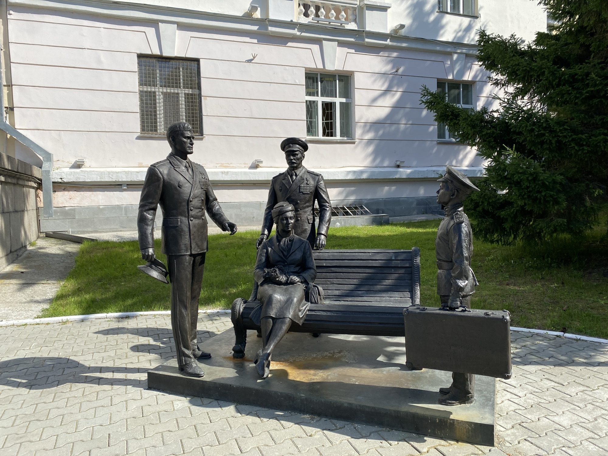 This is a bronze sculpture depicting three figures and a bench, situated in front of a light-colored building in Yekaterinburg, Russia. The sculpture is located near the Yekaterinburg Museum of History and Local Lore.
On the left, a man in a military uniform, holding a hat in his left hand, stands facing slightly right. In the center, a woman in a dress is seated on a dark bench, holding a bouquet of flowers. Behind her and slightly to her right stands another man in a military uniform, facing forward. To the right of the bench, a third man in a military uniform stands facing left, holding a briefcase. The figures are rendered with realistic detail. The environment consists of a paved walkway in the foreground, a patch of green grass behind the sculpture, and the facade of a building with windows in the background. A portion of a dark green coniferous tree is visible on the far right. The lighting suggests it is daytime, with shadows cast by the figures and the tree.