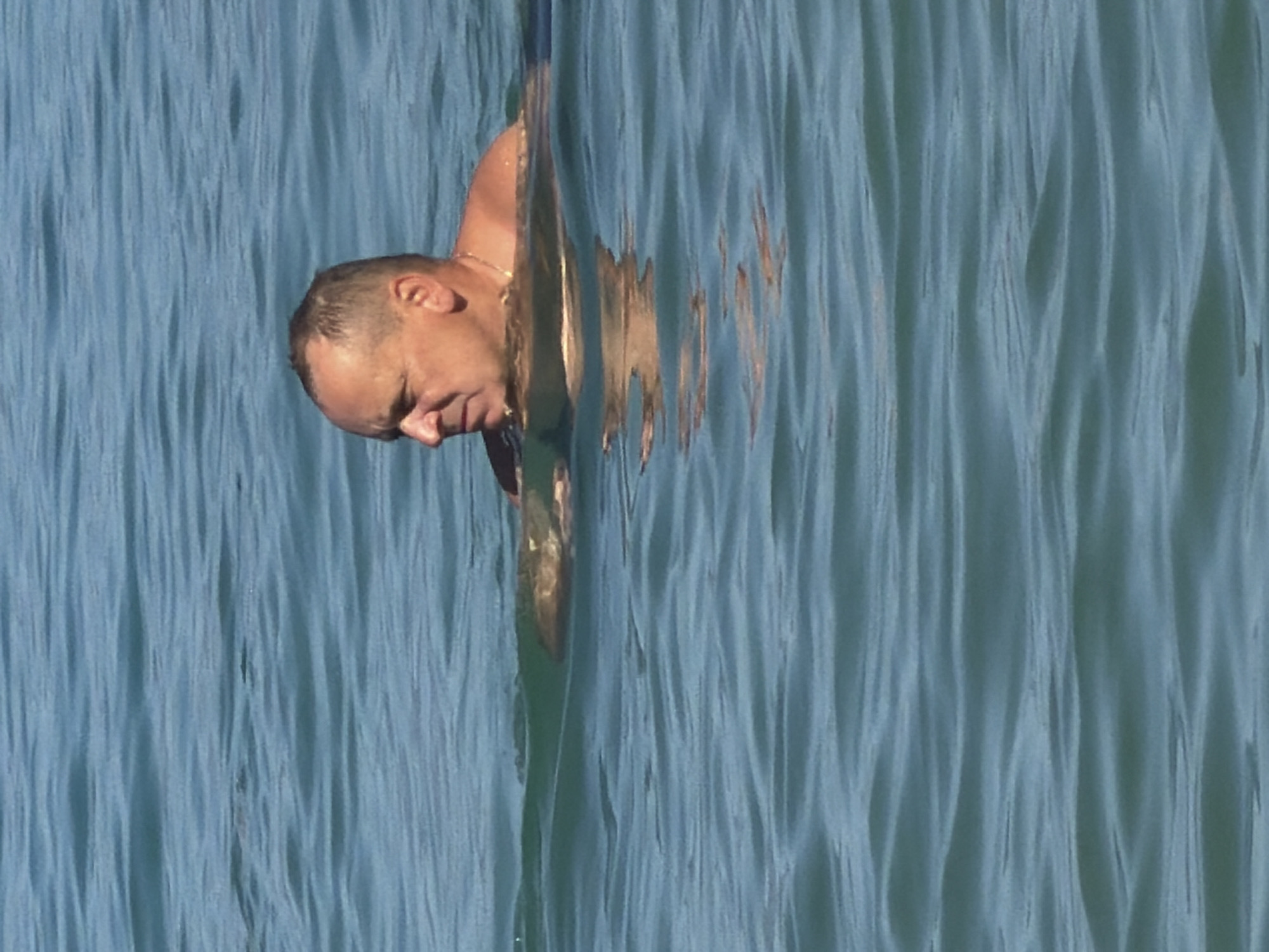 A man is swimming in the ocean, with his head and shoulders visible above the water. The water is a calm blue, with gentle ripples. The man has short, dark hair and a light complexion. He is wearing a gold chain around his neck. The image appears to be taken on a sunny day, with the sun reflecting off the water. The scene is likely taking place in the waters off the coast of Općina Baška, Croatia, a popular tourist destination known for its beautiful beaches and clear waters.