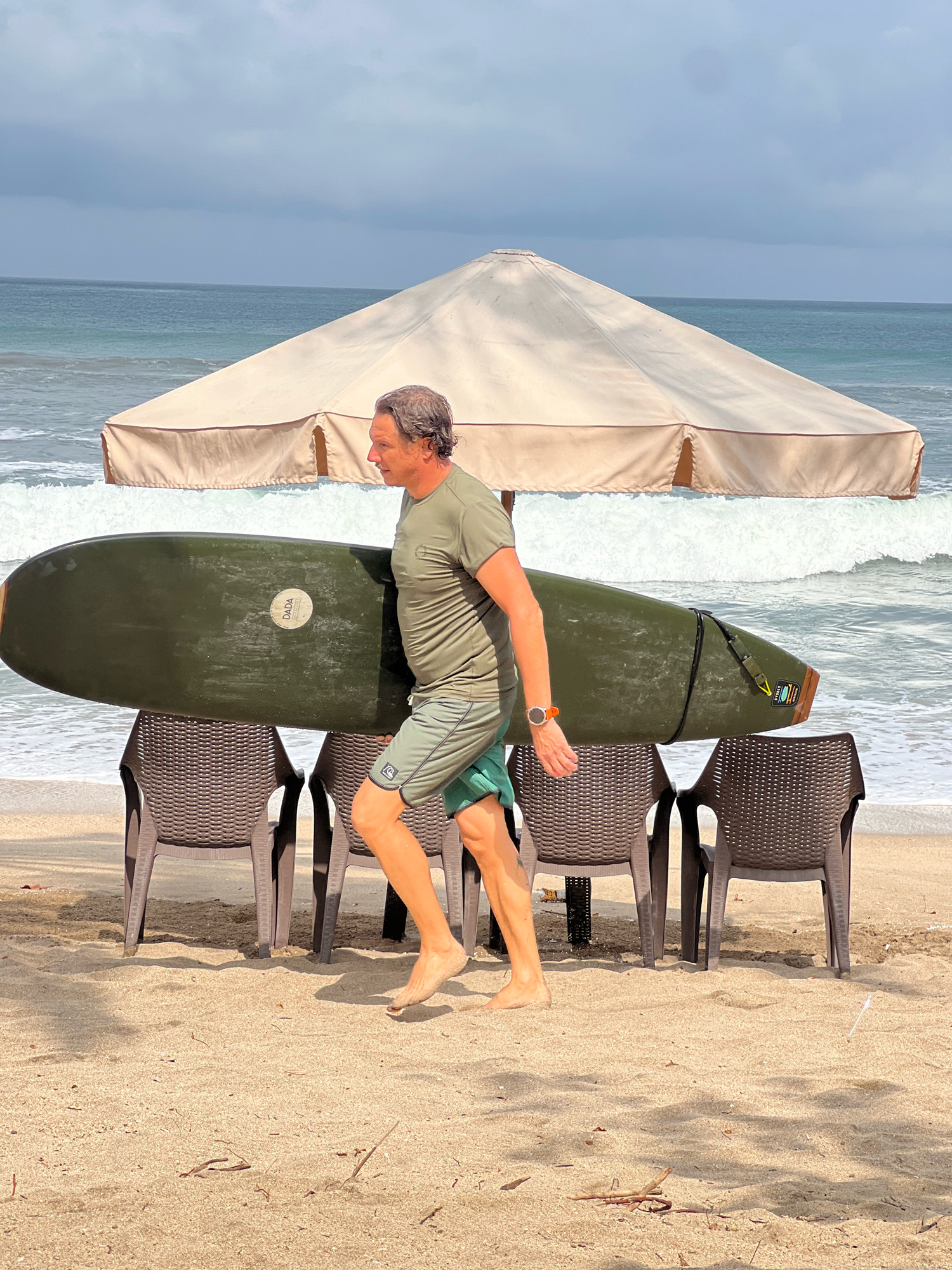 A man with graying hair, wearing a green t-shirt and green shorts, walks across a sandy beach in Kuta, Indonesia. He carries a dark green surfboard horizontally under his arm. In the background, a large beige umbrella provides shade over three dark brown plastic chairs. The ocean waves are visible behind the umbrella, with a cloudy sky overhead. The man appears to be heading towards the water, with his bare feet touching the sand. He is wearing an orange watch on his left wrist. The surfboard has a white circular logo with the word "DADA" on it, and a wooden tail.