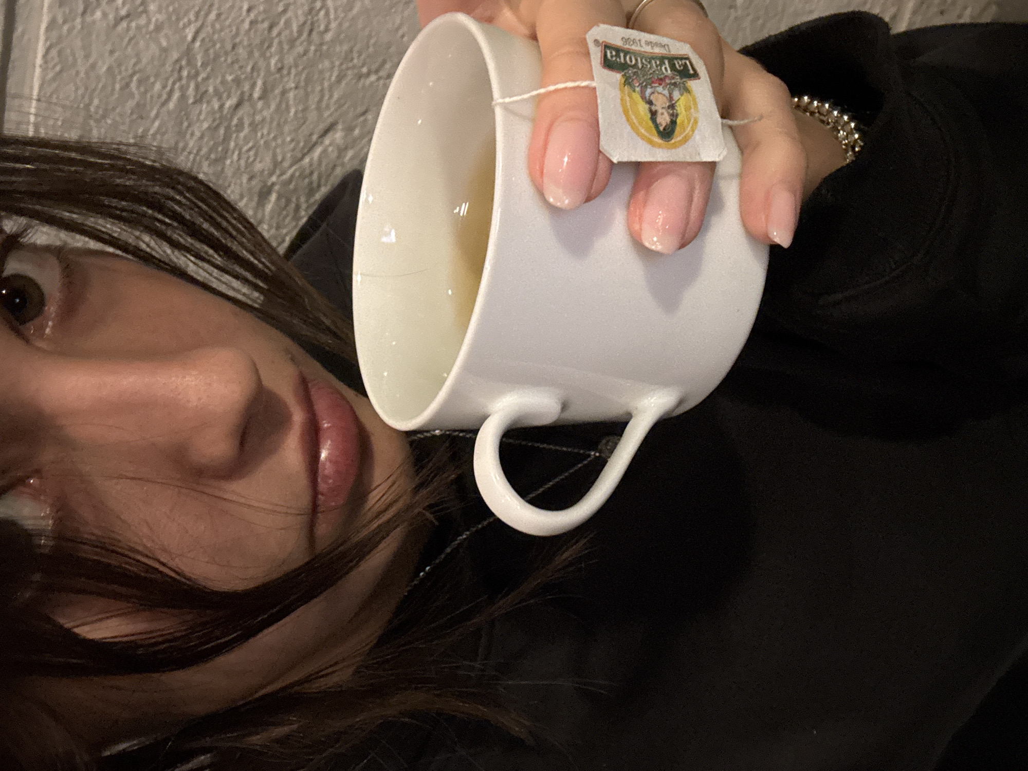 A close-up, eye-level shot captures a person, likely female, holding a white ceramic mug of tea. The subject's face is partially visible, showing one brown eye looking towards the camera, part of her nose, and glossy, plump lips. Dark hair frames her face, with strands falling across her cheek. She is wearing a dark, possibly black, long-sleeved garment, appearing to be a hoodie or sweatshirt, and a silver-colored beaded bracelet is visible on her wrist.
The hand holding the white mug features light pink/nude manicured nails. A tea bag tag, hanging from a string into the light-colored liquid, is prominently displayed. The tag clearly shows the brand name "La Pastora" and "Desde 1966" below an illustration of a woman. The tea appears to be steeping.
The background is a textured, light-colored wall, possibly stucco or concrete, suggesting an indoor setting. The casual composition, possibly a selfie taken from a reclining position, conveys a relaxed and intimate mood. The tea brand "La Pastora" provides a cultural cue consistent with the location context of Mexico City, Mexico.