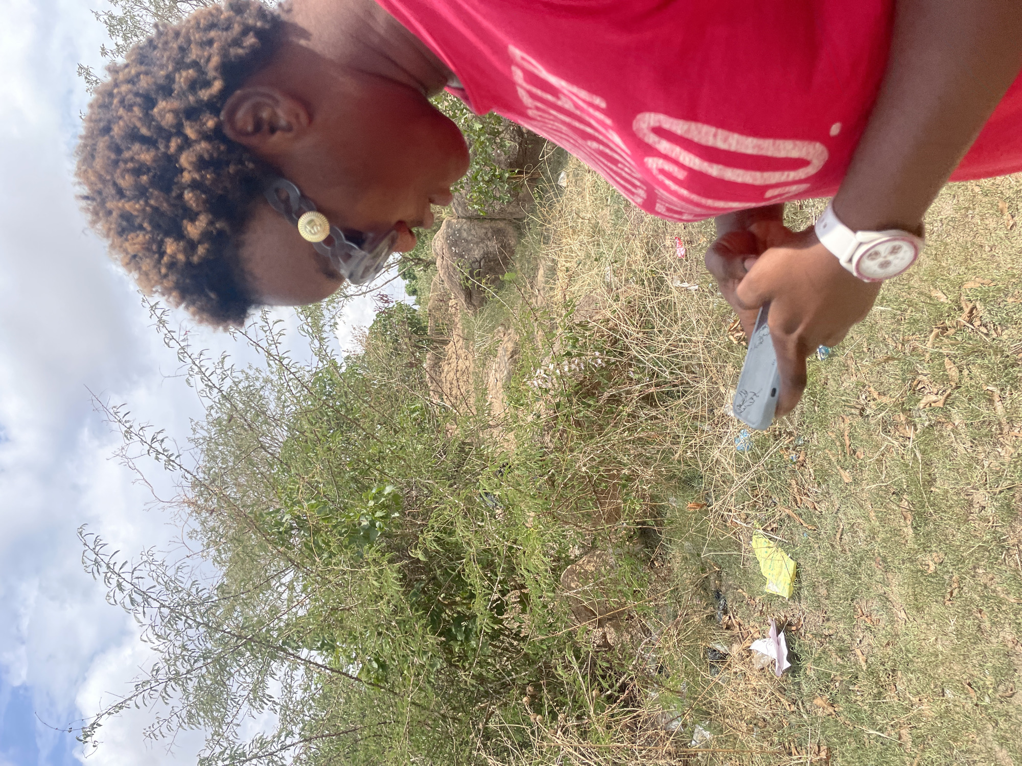 The image depicts a woman outdoors, likely in a natural or semi-natural environment in Jos, Nigeria. She is wearing a red t-shirt and a white watch. She is holding a gray object in her hand. The surrounding environment consists of grass, sparse vegetation, and a sky with light clouds. Trash is scattered on the ground. A rock can be seen behind the trees.
