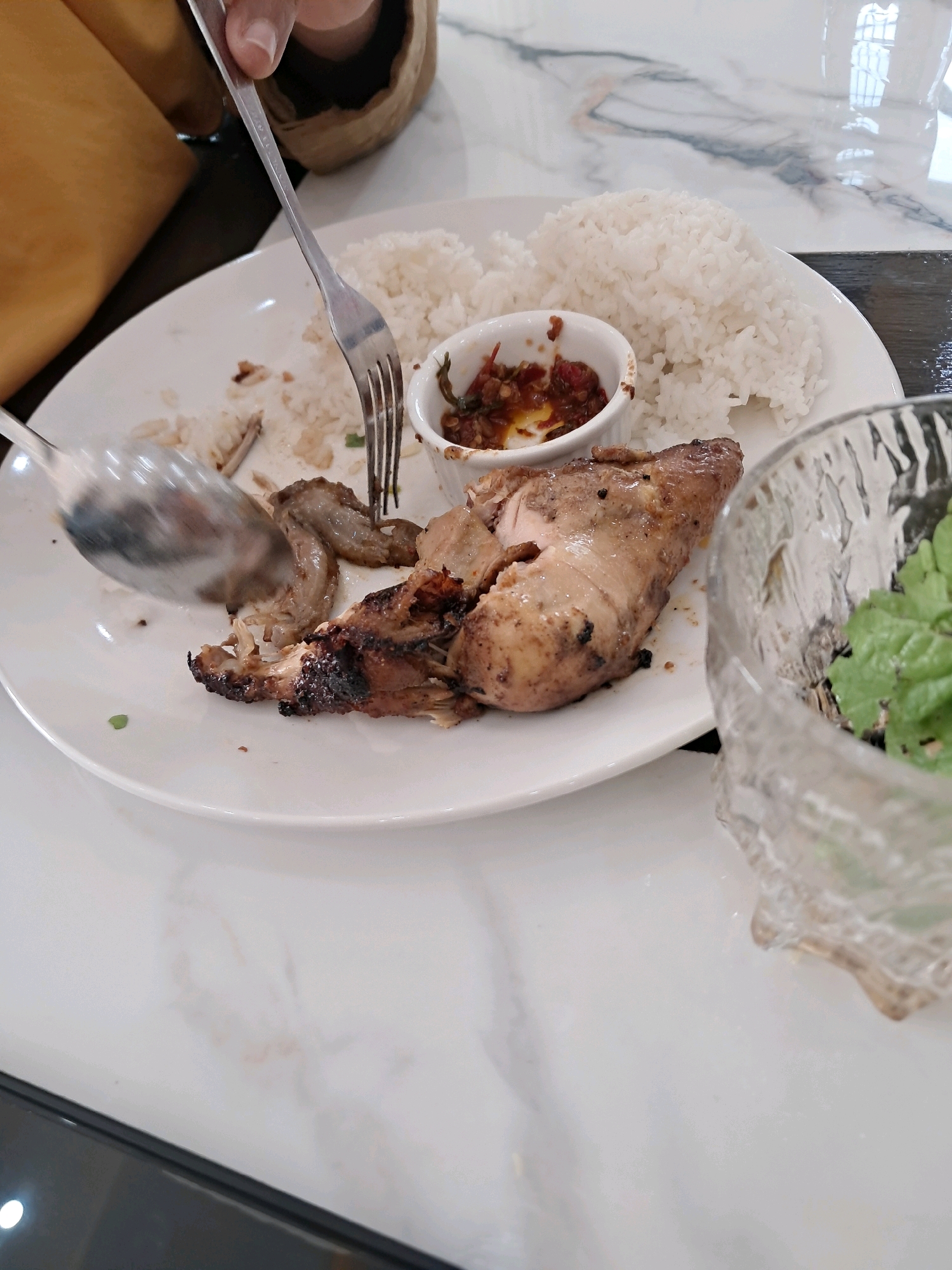 A partially eaten meal of grilled chicken and rice is on a white plate. A spoon is seen in the process of scooping up a piece of chicken. A small bowl of chili sauce is also on the plate, as is a glass bowl containing green leafy vegetables. The scene takes place at a restaurant in Mempawah, Indonesia, with a marble-patterned table and a blurred background showing other diners.