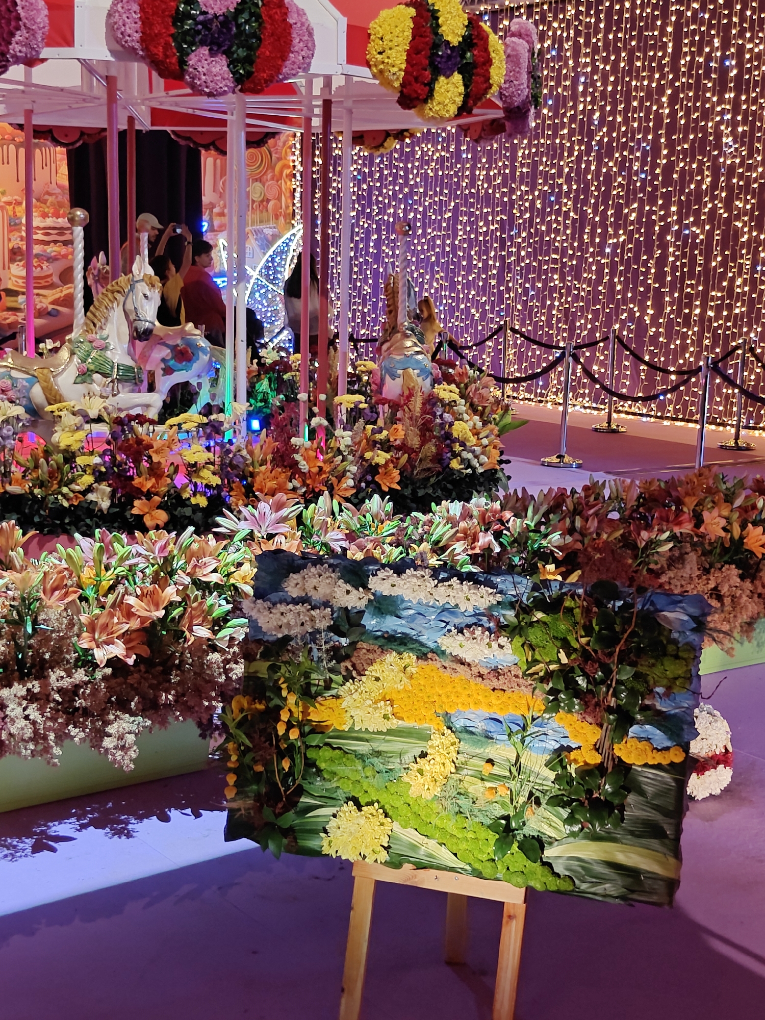 The image depicts an indoor scene, likely an exhibition or event space, featuring a carousel and floral arrangements. A white carousel horse is visible in the foreground, adorned with floral decorations. Behind it, a backdrop of numerous small, twinkling lights creates a luminous effect. Several people are present, some of whom appear to be taking photographs. The environment is decorated with abundant, colorful floral displays in shades of orange, yellow, pink, and red. In the center foreground, a large, three-dimensional artwork, possibly a landscape, is displayed on a wooden easel. The artwork is constructed using various materials, including what appear to be small flowers or textured elements, to depict a scene with water, greenery, and yellow and orange elements. The floor is a reflective, purple-toned surface, suggesting artificial lighting. The overall impression is one of a festive and decorative setting. The provided context of Tel-Aviv, Israel, suggests this may be an event or exhibition held within the city, though no specific landmarks or identifying features of Tel-Aviv are discernible in the image itself.