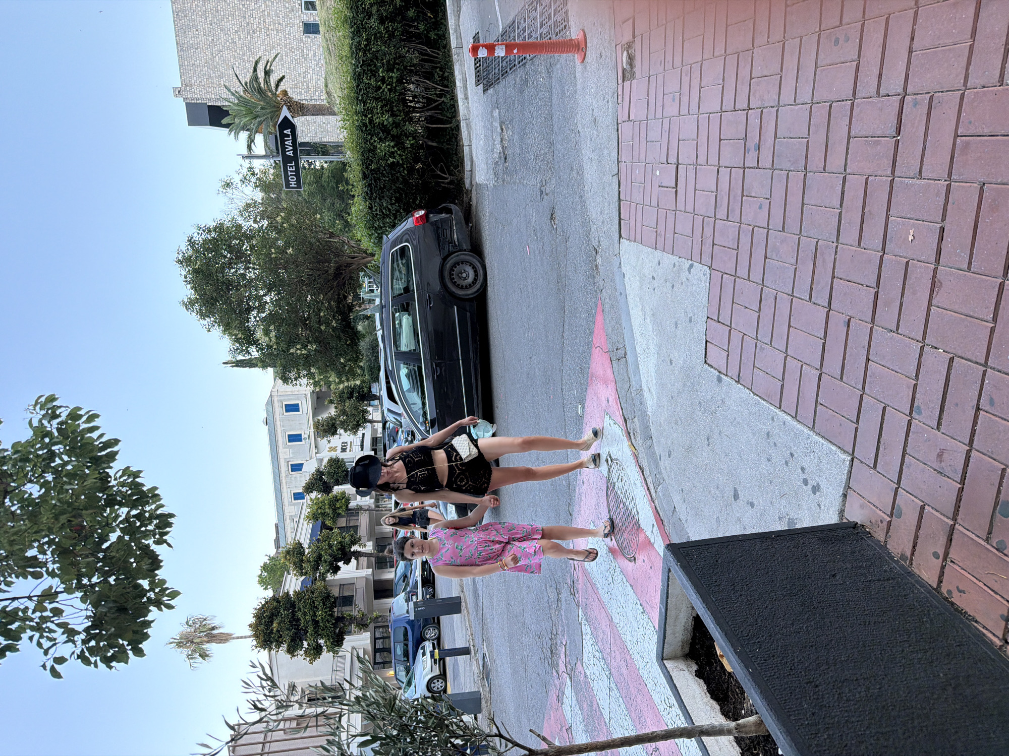 In Budva, Montenegro, a woman and a young girl are crossing a pedestrian street. The woman, dressed in a black lace outfit and a wide-brimmed hat, holds the girl's hand. The girl wears a pink floral dress. They are walking on a crosswalk with red and white stripes. In the background, cars are parked on the side of the street, and a sign pointing to "HOTEL AVALA" is visible. Trees and buildings line the street, under a clear blue sky. The scene appears to be a street in the city center.