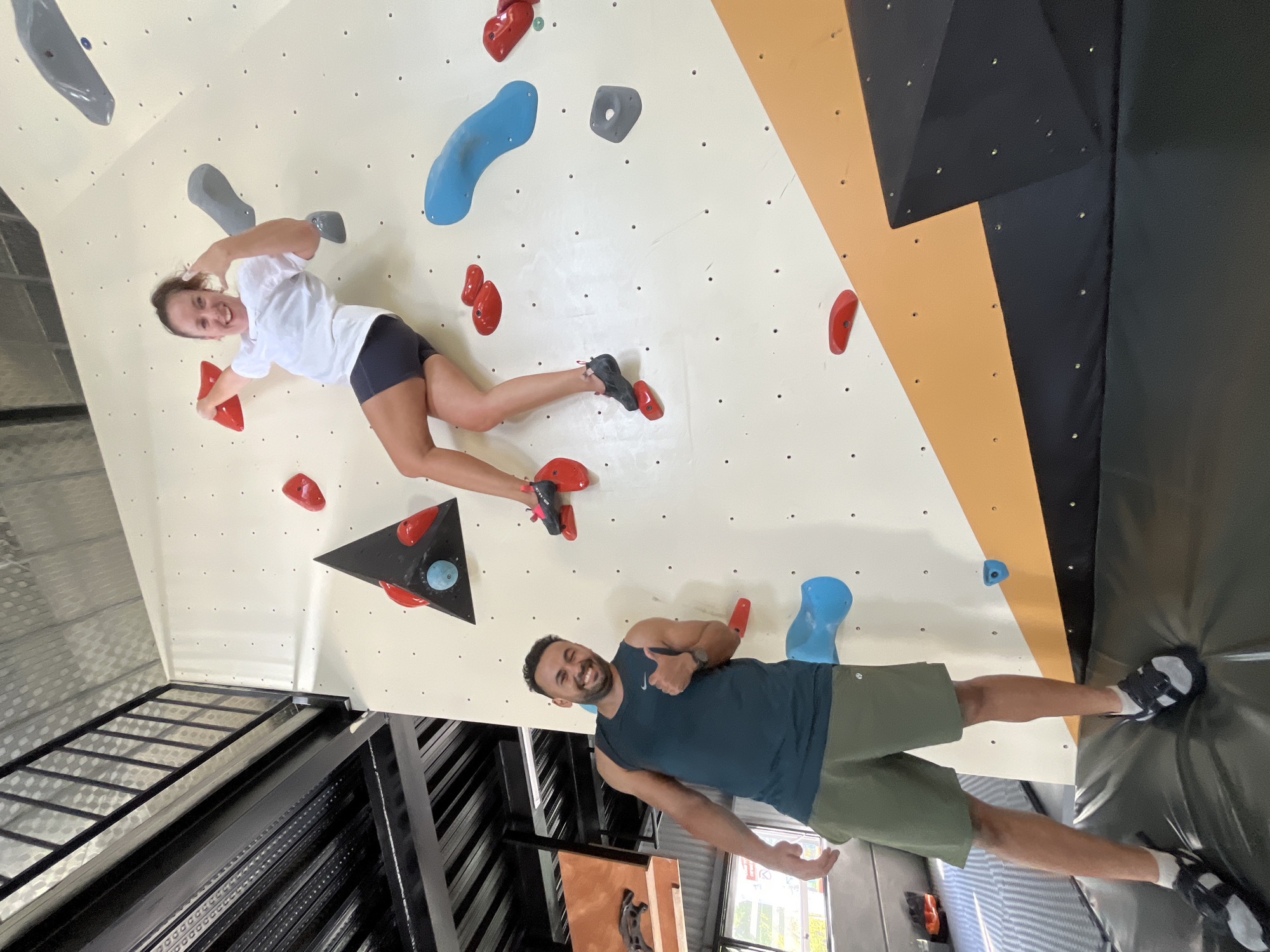 This image depicts an indoor bouldering gym located in the city of Đà Nẵng, Vietnam. The scene features two individuals. On the upper right, a female with dark hair pulled into a ponytail, wearing a white short-sleeved t-shirt and dark athletic shorts, is actively climbing an off-white bouldering wall. Her right hand grips a red climbing hold, while her left hand is raised near her face, forming a celebratory gesture as she smiles broadly towards the camera. She is wearing black climbing shoes with pink accents, with her feet positioned on red holds.
Below her, towards the lower left, a male with short dark hair and a beard stands on a black crash mat at the base of the climbing wall. He wears a dark teal sleeveless athletic tank top featuring a white Nike swoosh logo on the left chest and olive green athletic shorts with a white Lululemon logo on the left thigh. He is smiling widely at the camera, giving a thumbs-up gesture with his right hand. He wears black sandals over white socks.
The climbing wall is predominantly off-white, marked with numerous small black attachment points. Various climbing holds in red, blue, and gray are distributed across its surface. A prominent black triangular volume, equipped with additional red and a light blue hold, is positioned in the upper central area of the wall. The lower right section of the wall features a large orange diagonal panel above a black base.
The background in the upper left reveals an industrial-style ceiling and upper level, characterized by black metal beams, a black metal railing, and corrugated silver ceiling panels. Fluorescent lighting fixtures are visible on the ceiling structure. To the left of the male, a partial view of another section of the wall with a brown panel and a dark climbing hold is visible, with an out-of-focus exterior view beyond. The floor area beneath the climbing wall is covered with black crash mats.