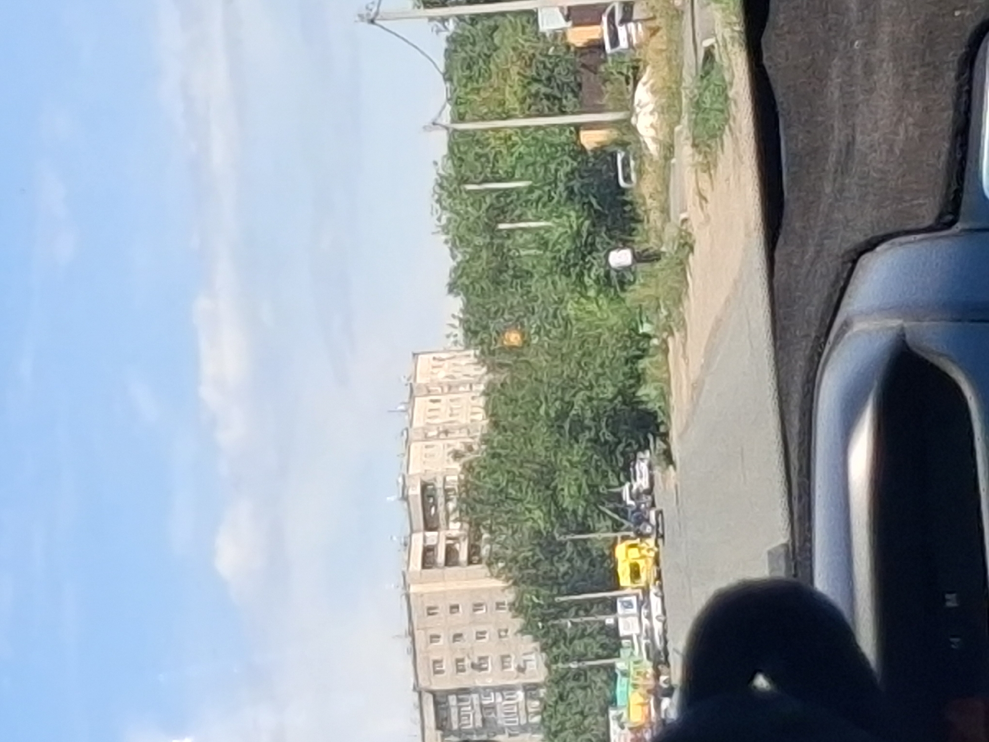 The image shows a street scene in Semey, Kazakhstan, on a sunny day with a partly cloudy sky. In the background, a multi-story apartment building with a light-colored facade stands behind a dense line of green trees. The foreground is a street with several vehicles, including a yellow truck, white cars, and a silver car. Some of the vehicles are parked along the side of the road, while others appear to be moving or stopped in traffic. There are also utility poles and wires visible. The street is paved, and there is some vegetation growing along the sides. In the middle ground, a person in a white shirt and dark pants is standing near the trees. The scene appears to be a typical urban environment in Semey.