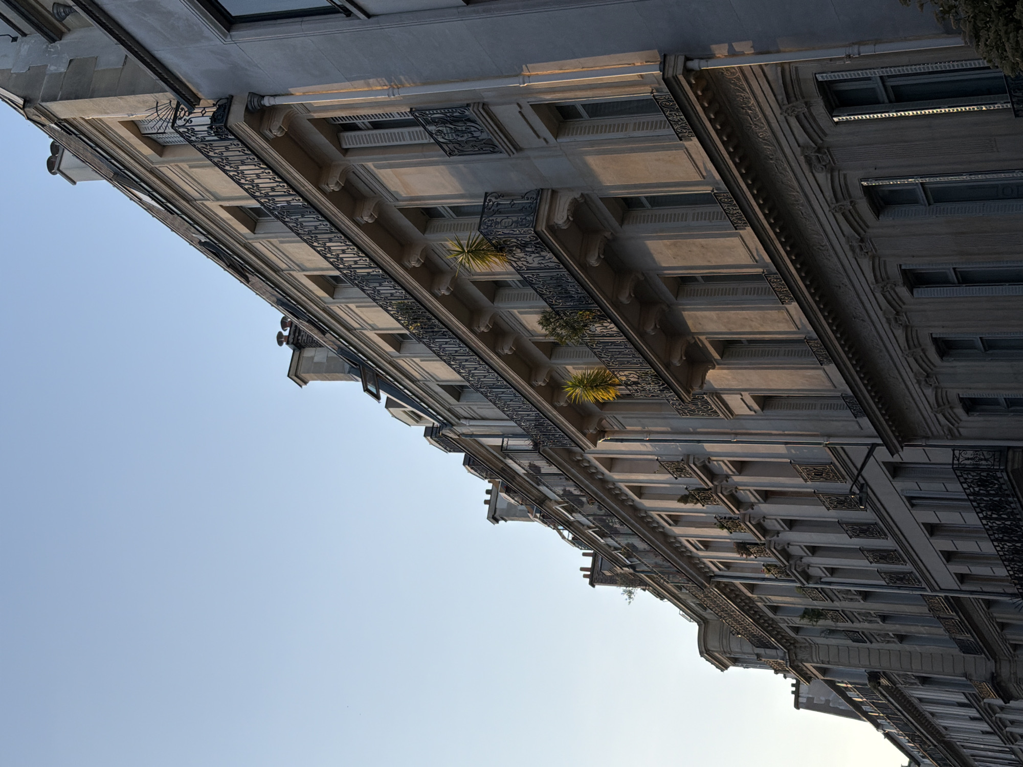 The image captures a dramatic, low-angle view of the ornate facades of Parisian buildings, stretching diagonally across the frame against a clear, light blue sky. The perspective looks sharply upwards, emphasizing the height and intricate details of the architecture.
The primary subjects are the multi-storied stone buildings, characterized by their classic Haussmannian style. They feature numerous windows and balconies adorned with elaborate, dark wrought-iron railings and grilles. These balconies are a prominent feature, showcasing intricate scrollwork and patterns. Several balconies hold potted plants or small trees, adding patches of green to the stone edifice.
Notable architectural details include decorative stone carvings such as corbels beneath the balconies, pronounced cornices, and detailed moldings that delineate each floor. The building materials appear to be light-colored stone or stucco, possibly limestone, which is subtly shadowed and highlighted by the natural light.
The lighting suggests late afternoon or early evening, with warm, directional light catching the upper sections and projecting long shadows that define the architectural relief. This creates depth and texture across the facades. The sky is a uniform, pale blue, devoid of clouds, occupying the lower and left portions of the image.
There are no people or discernible activity, presenting a static, architectural study. No visible text can be identified on the buildings. The image conveys a sense of grandeur and elegance, highlighting the timeless beauty of Parisian architecture through an unusual and dynamic composition.