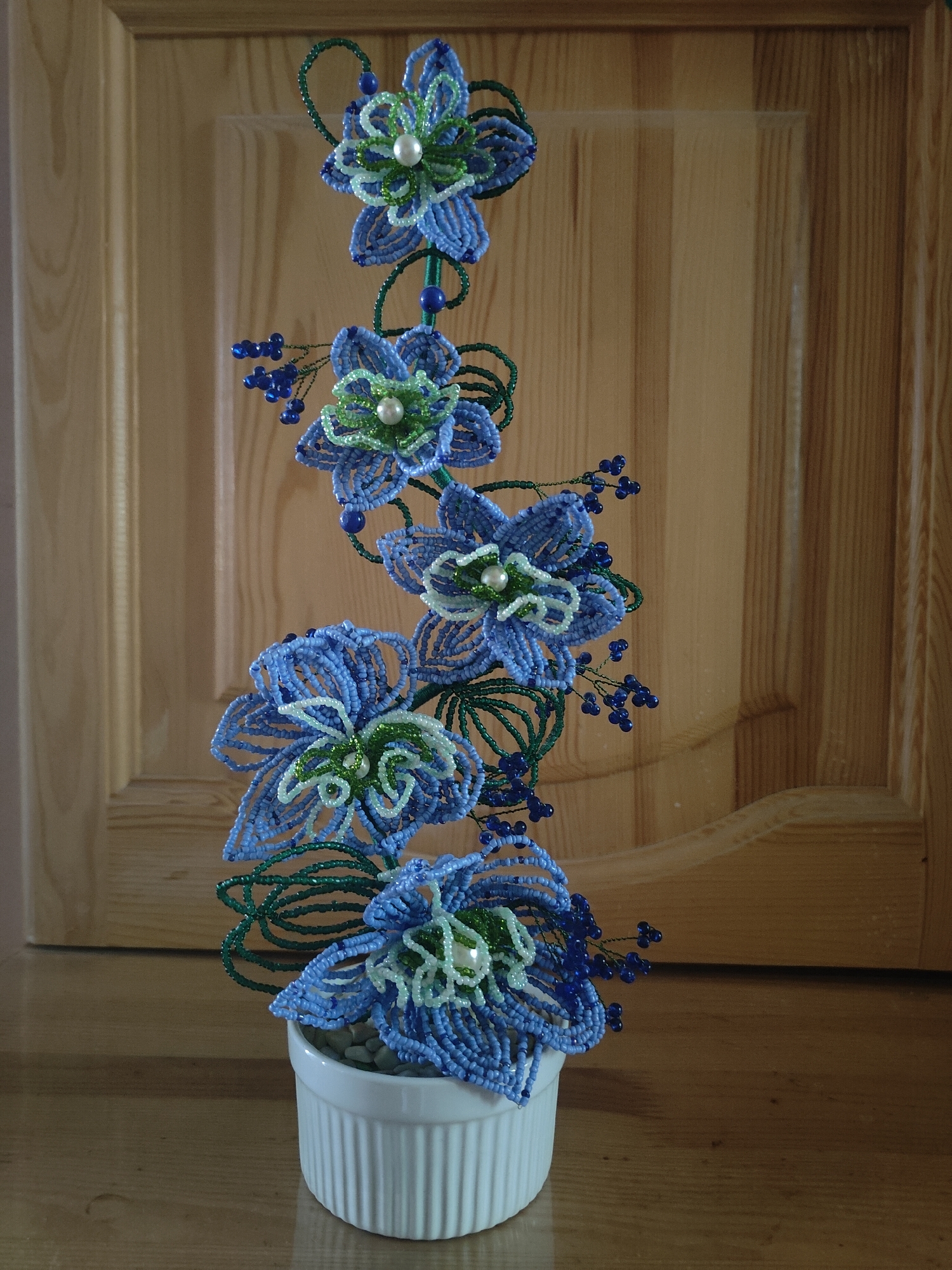 A beaded floral arrangement featuring blue and green flowers with pearl centers stands in a white ribbed ceramic pot. The arrangement is positioned in front of a light brown wooden paneled door. The flowers are intricate, with multiple delicate petals made of small beads. Green beaded vines and small clusters of dark blue beads resembling berries are interspersed among the flowers. The pot is filled with small, light-colored pebbles. The lighting suggests an indoor environment, possibly a home, with the natural wood grain of the door providing a warm backdrop.